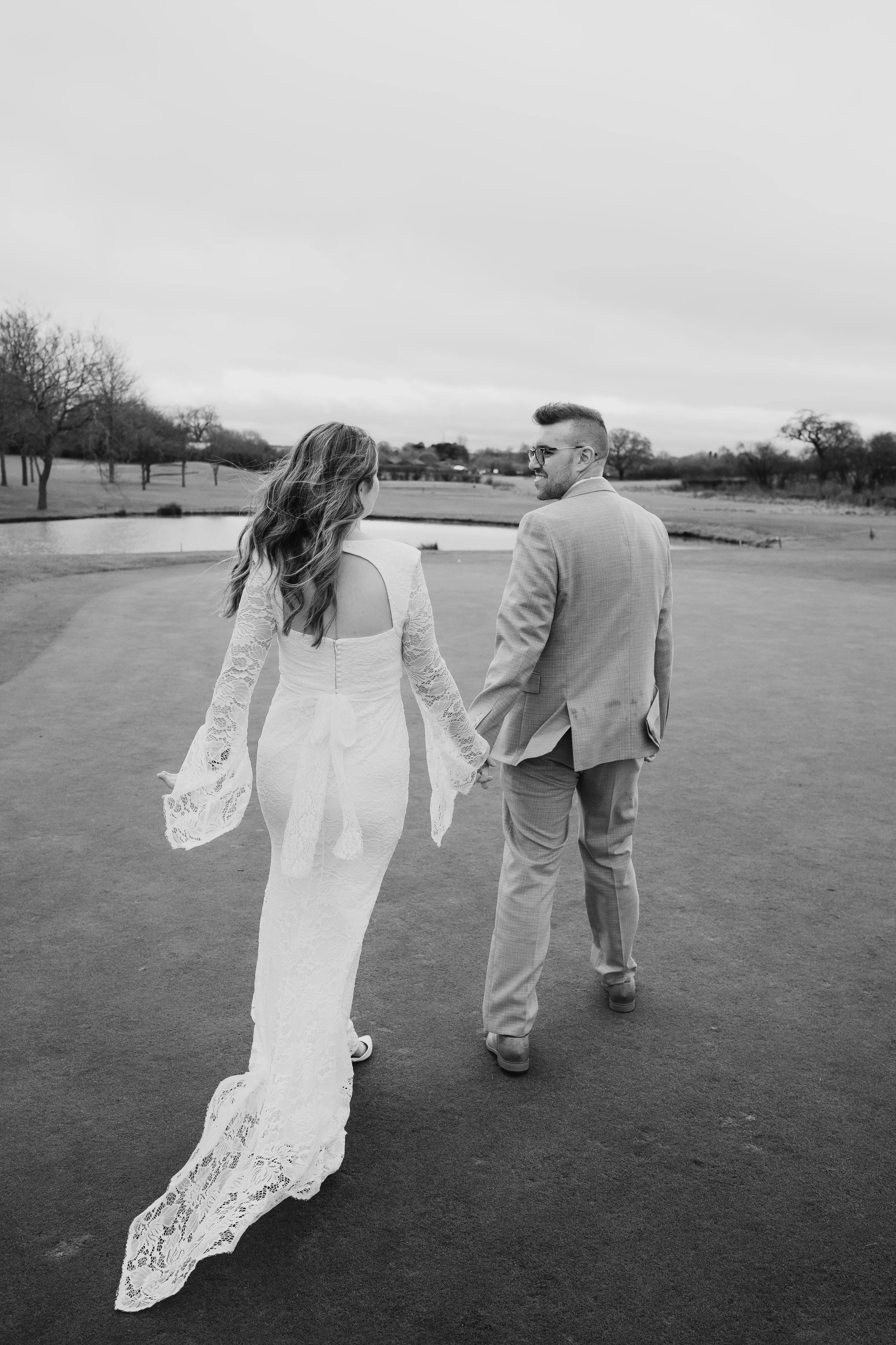 A black and white photo of a bride and groom walking hand in hand on a golf course, with trees and a pond in the background.