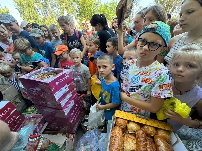 A group of children and adults gathered outdoors around a table with boxes of food and baked goods. The people are looking at the food, some holding bags. Trees are visible in the background.
