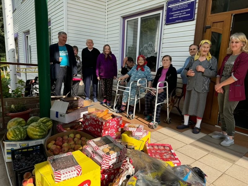 A group of people standing outside a building with fresh produce and packaged food items on the ground in front of them. One person uses a walker.