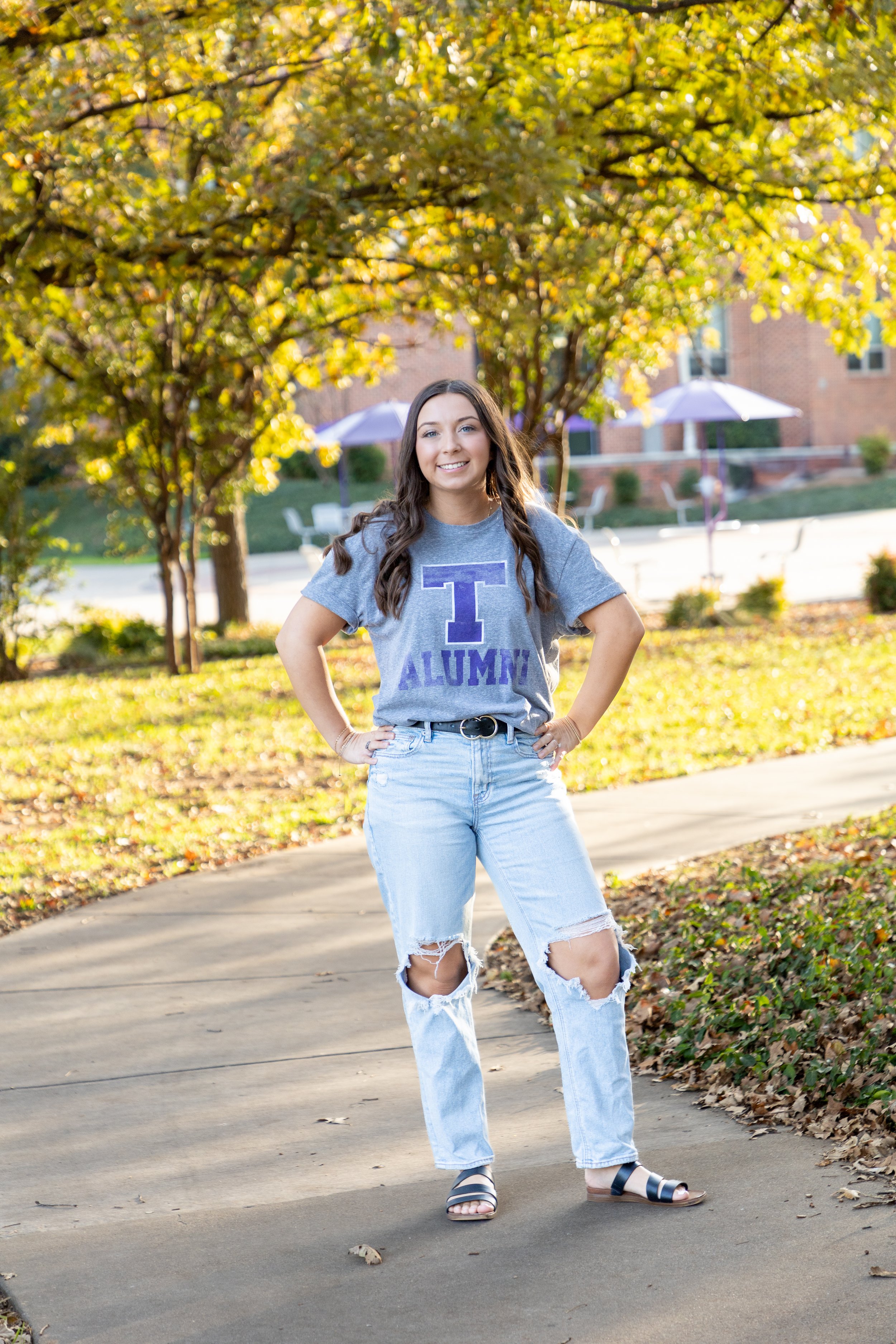 A young woman with long brown hair, smiling, standing outdoors on a curved concrete path, wearing a gray T-shirt with purple lettering that says "T ALUM" and ripped light blue jeans, with trees and umbrellas in the background.
