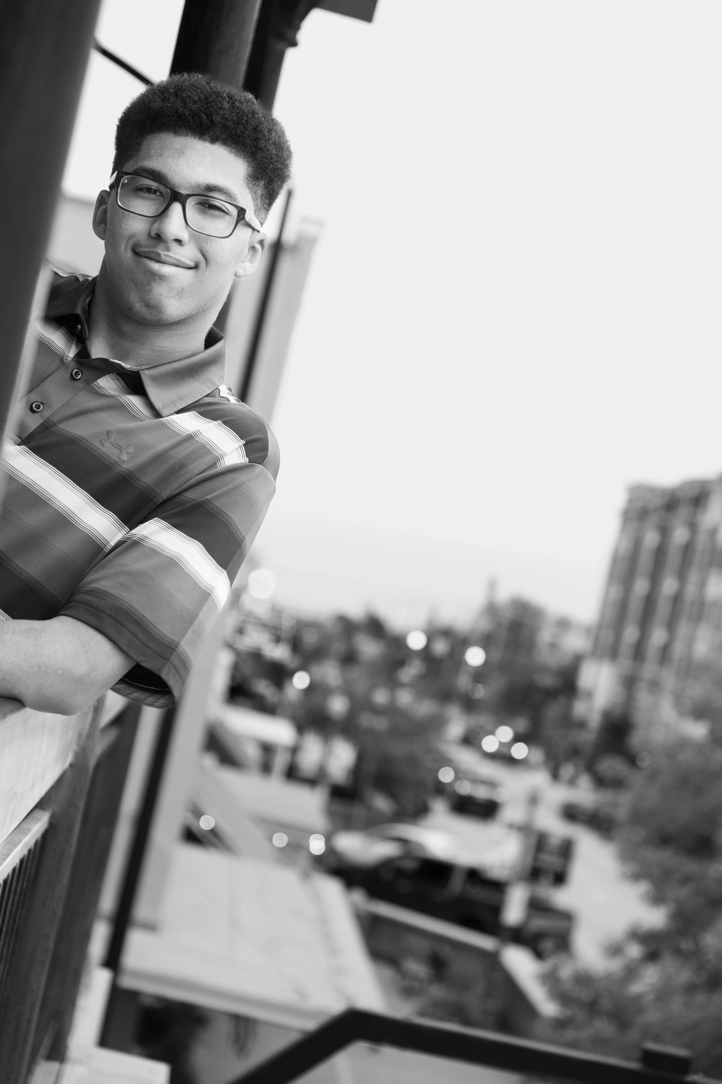 A young man with glasses and short curly hair, smiling softly, standing on a balcony or ledge with a cityscape in the background. The photo is in black and white.