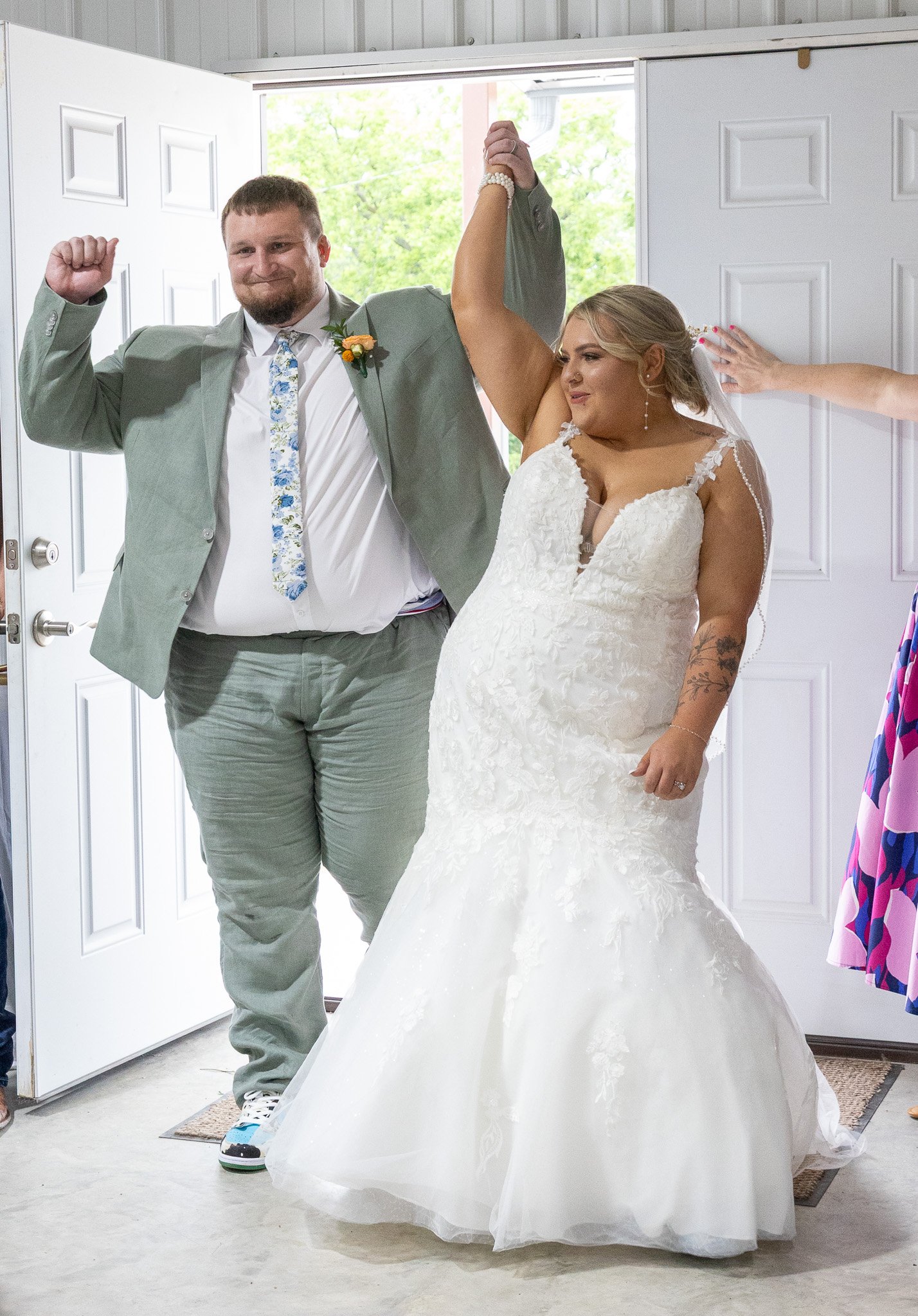 A bride and groom are celebrating, with the groom in a gray suit and the bride in a white wedding gown. The groom is raising his fist, and the bride is raising her arm in a victorious pose, inside a home with an open door and greenery outside.