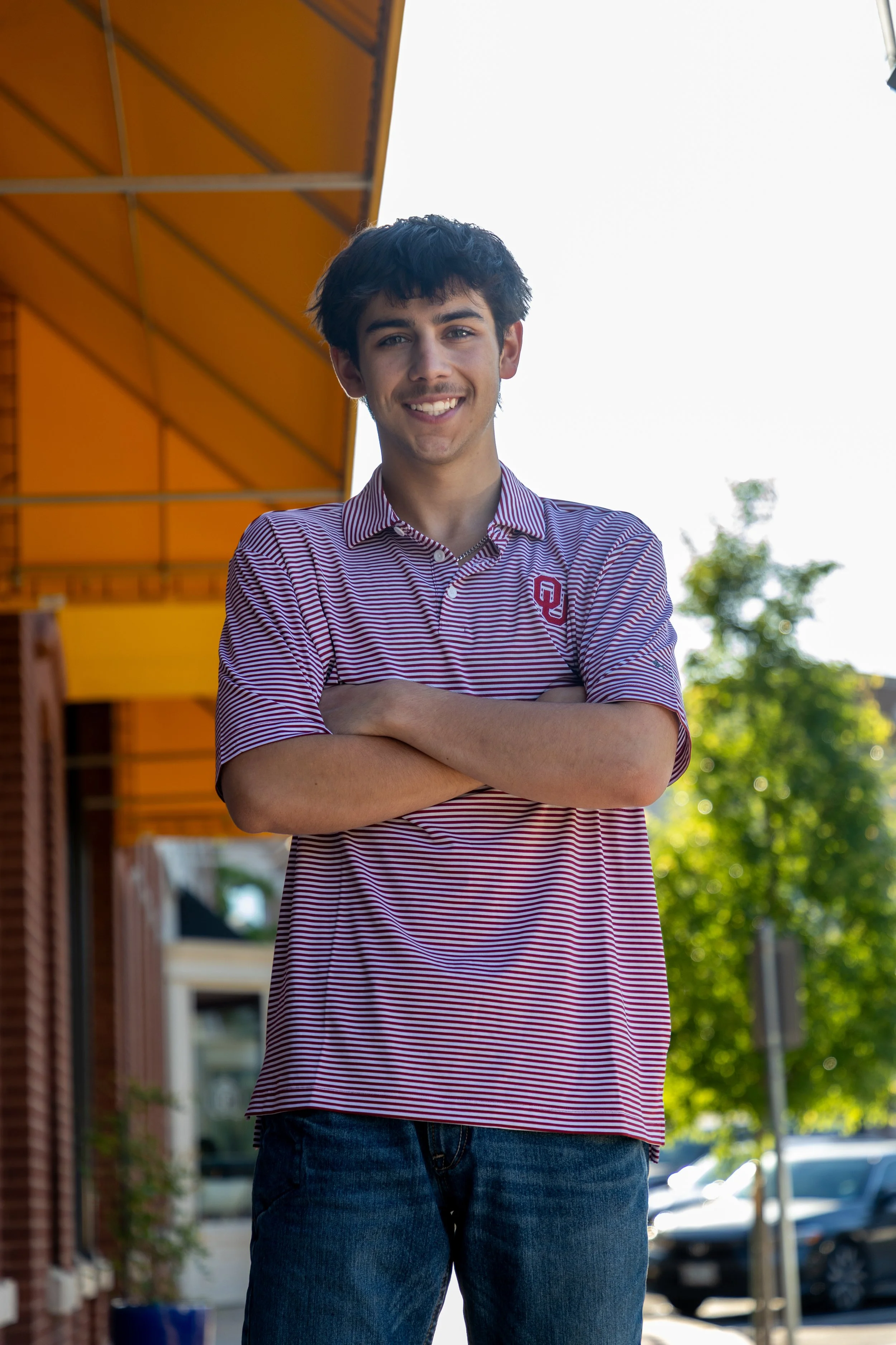 A young man with dark hair and light skin smiling, standing outdoors with arms crossed, wearing a striped red and white polo shirt with an OU logo, and dark jeans, in front of a building with orange awning and greenery.