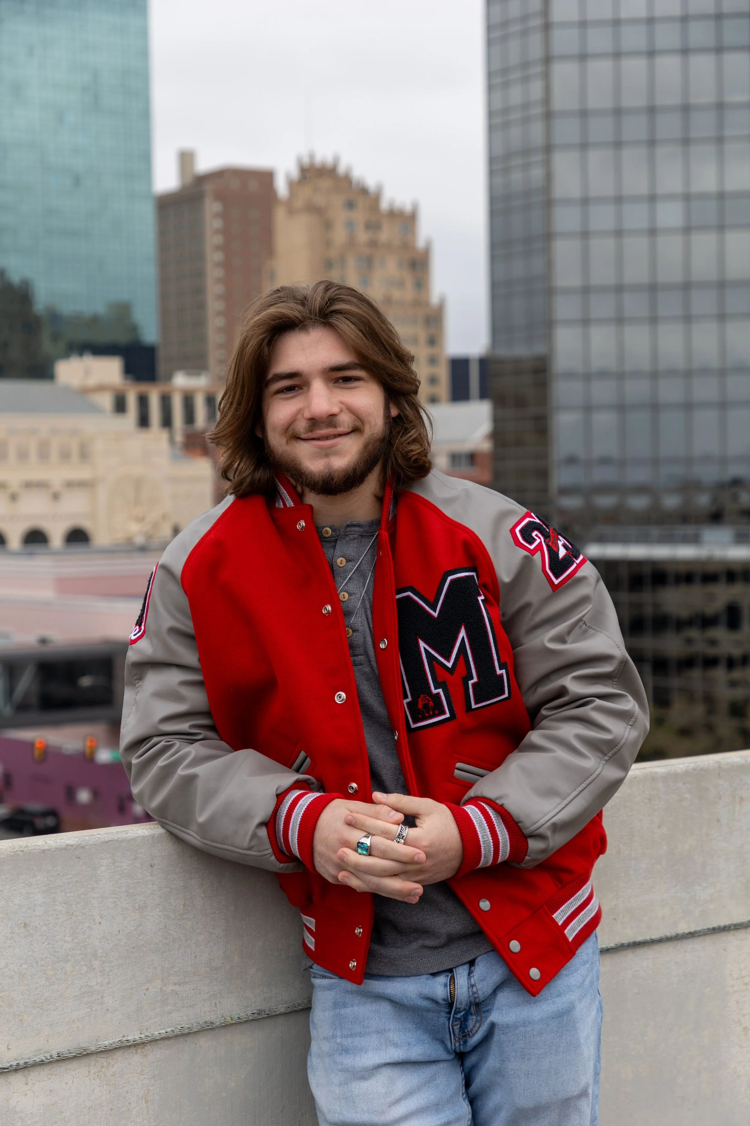 A young man with long hair and a beard standing outdoors on a rooftop with city buildings in the background. He is wearing a red and gray letterman jacket.