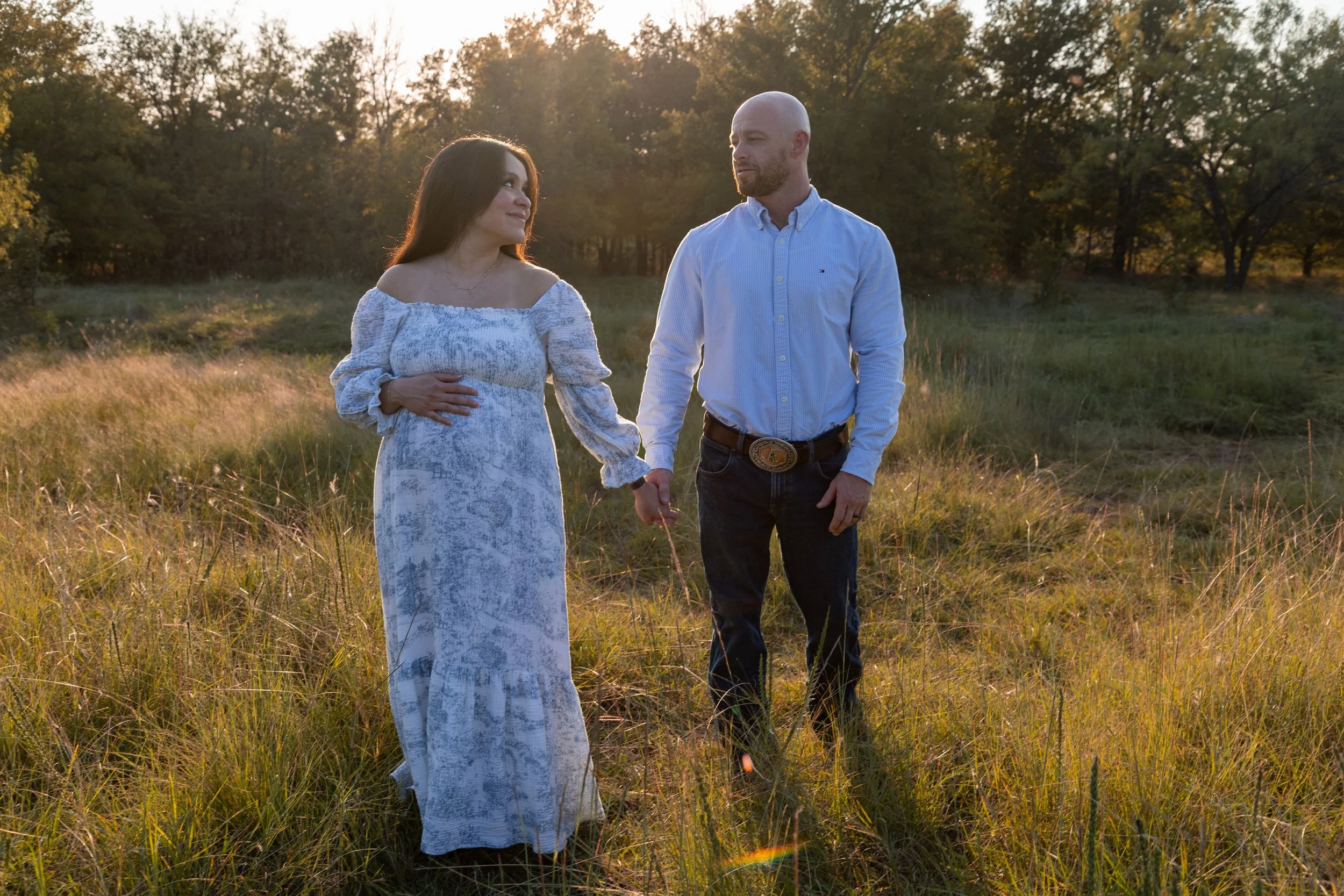 A pregnant woman and a man holding hands in a grassy field during sunset, with trees in the background.