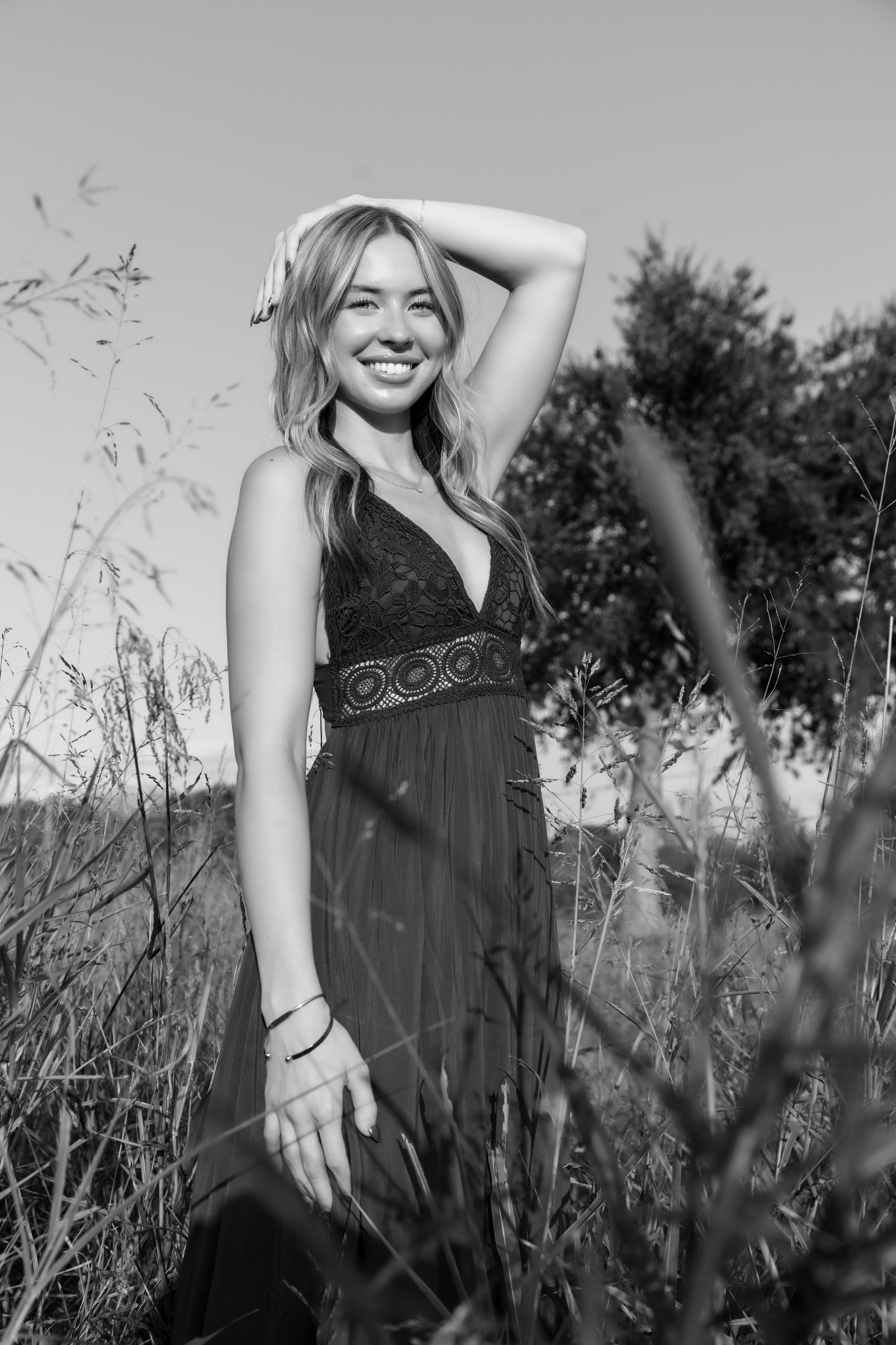 A smiling woman in a dress standing in a field of tall grass with trees in the background, captured in black and white.
