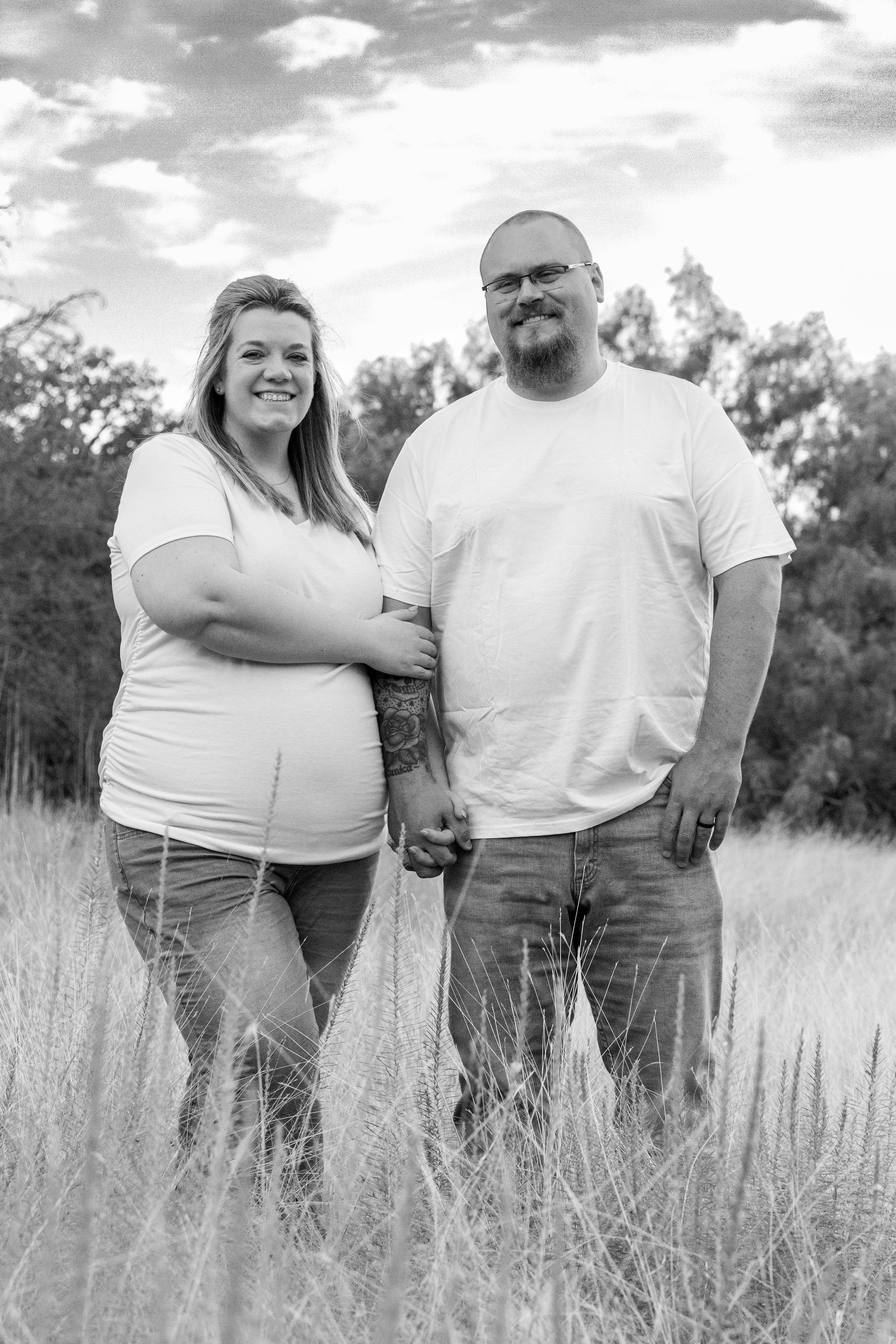 A black and white photo of a couple holding hands and smiling outdoors in a field of tall grass, with trees and a partly cloudy sky in the background.