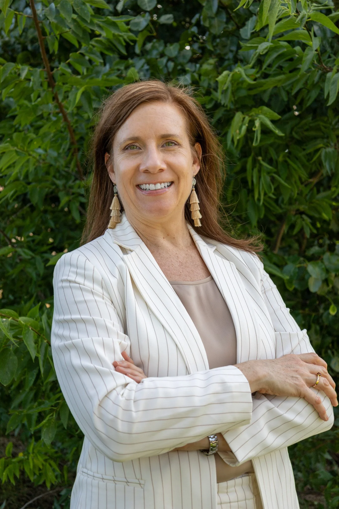 A woman with red hair wearing a white striped blazer and tiered earrings, standing outdoors with green foliage in the background.