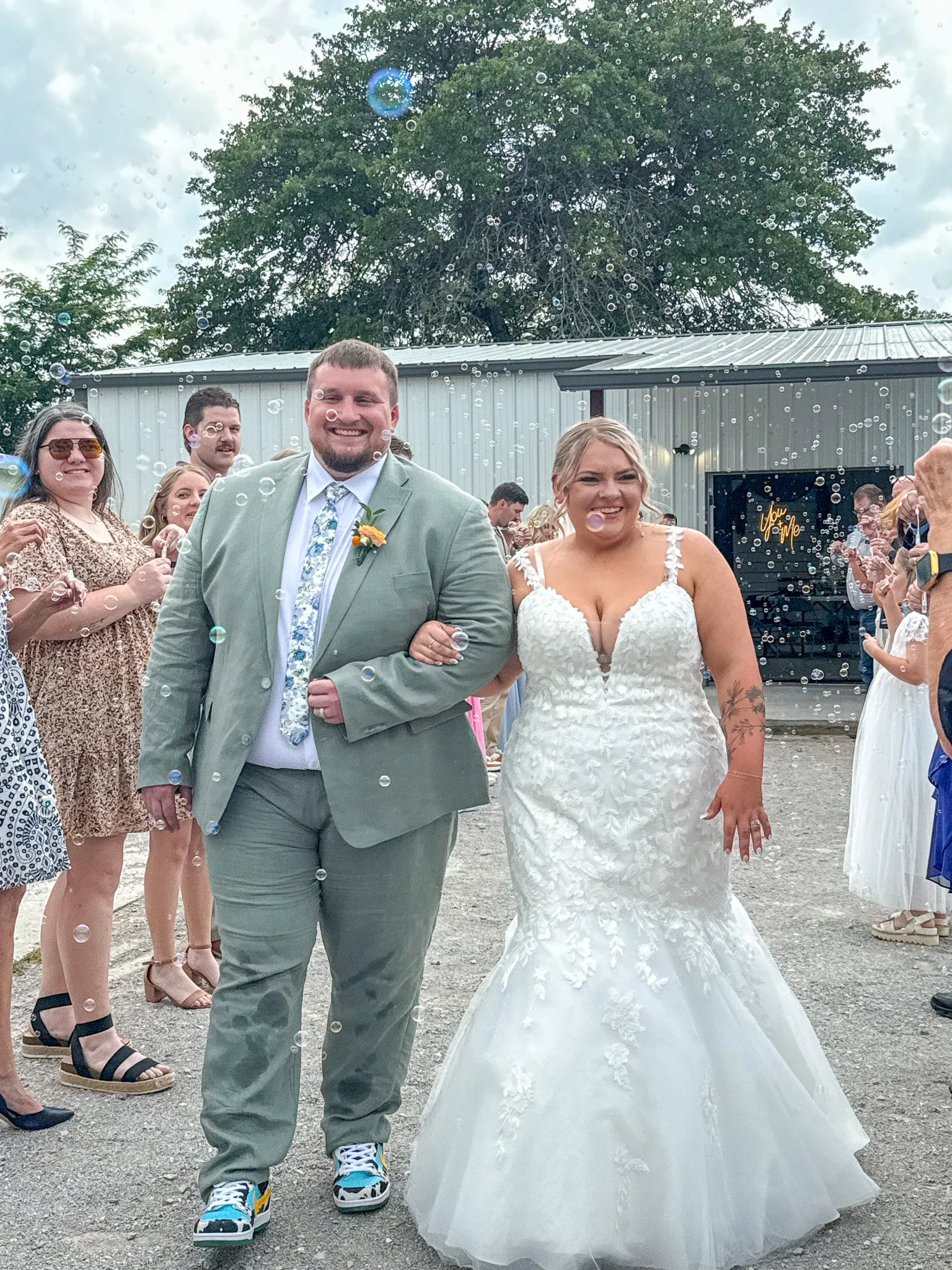 Bride and groom walking outside, surrounded by friends and family, during a wedding celebration, with bubbles and a cloudy sky.