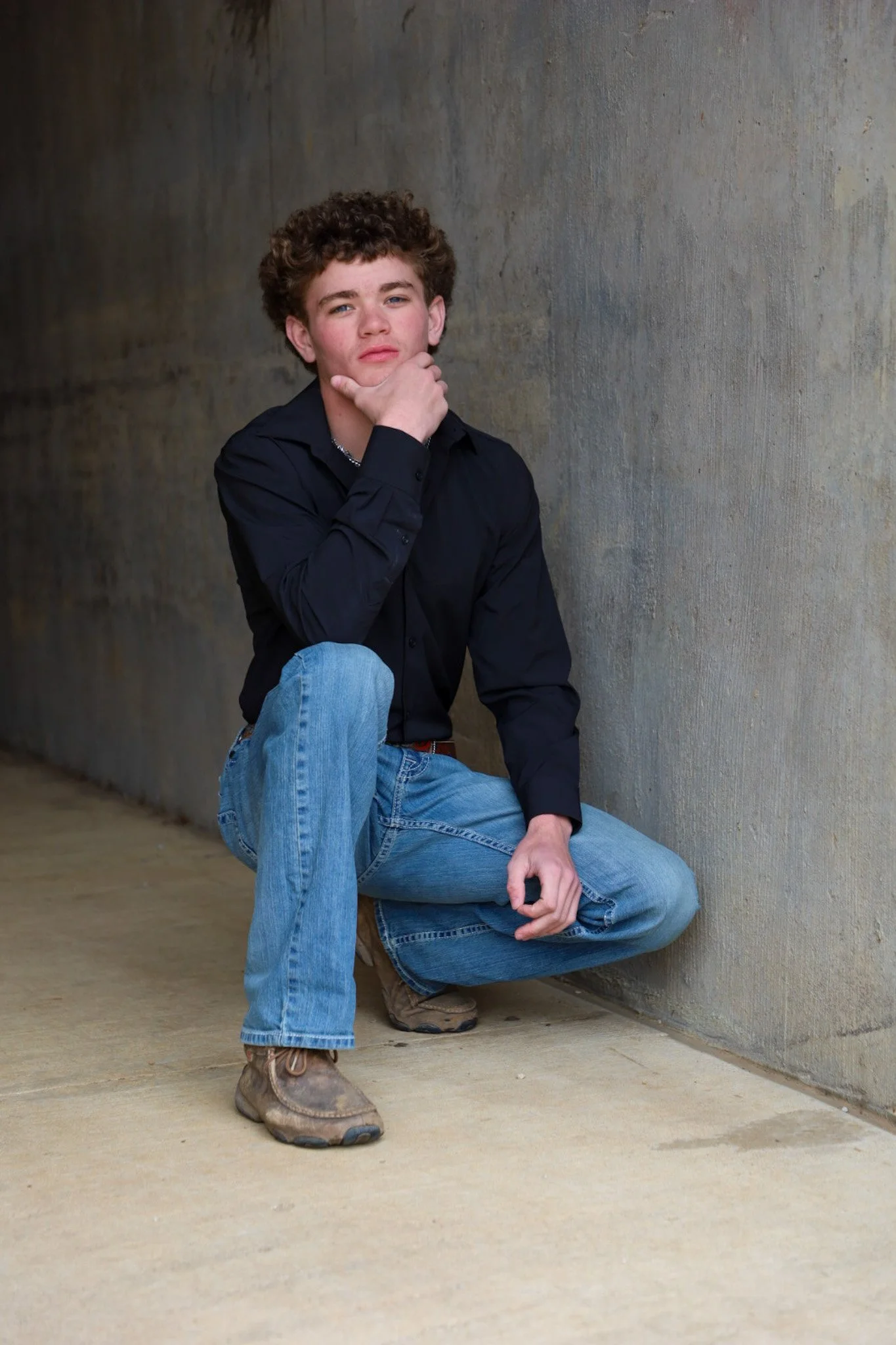 Young man in black shirt and blue jeans crouching against a concrete wall, with hand on chin in a thoughtful pose.