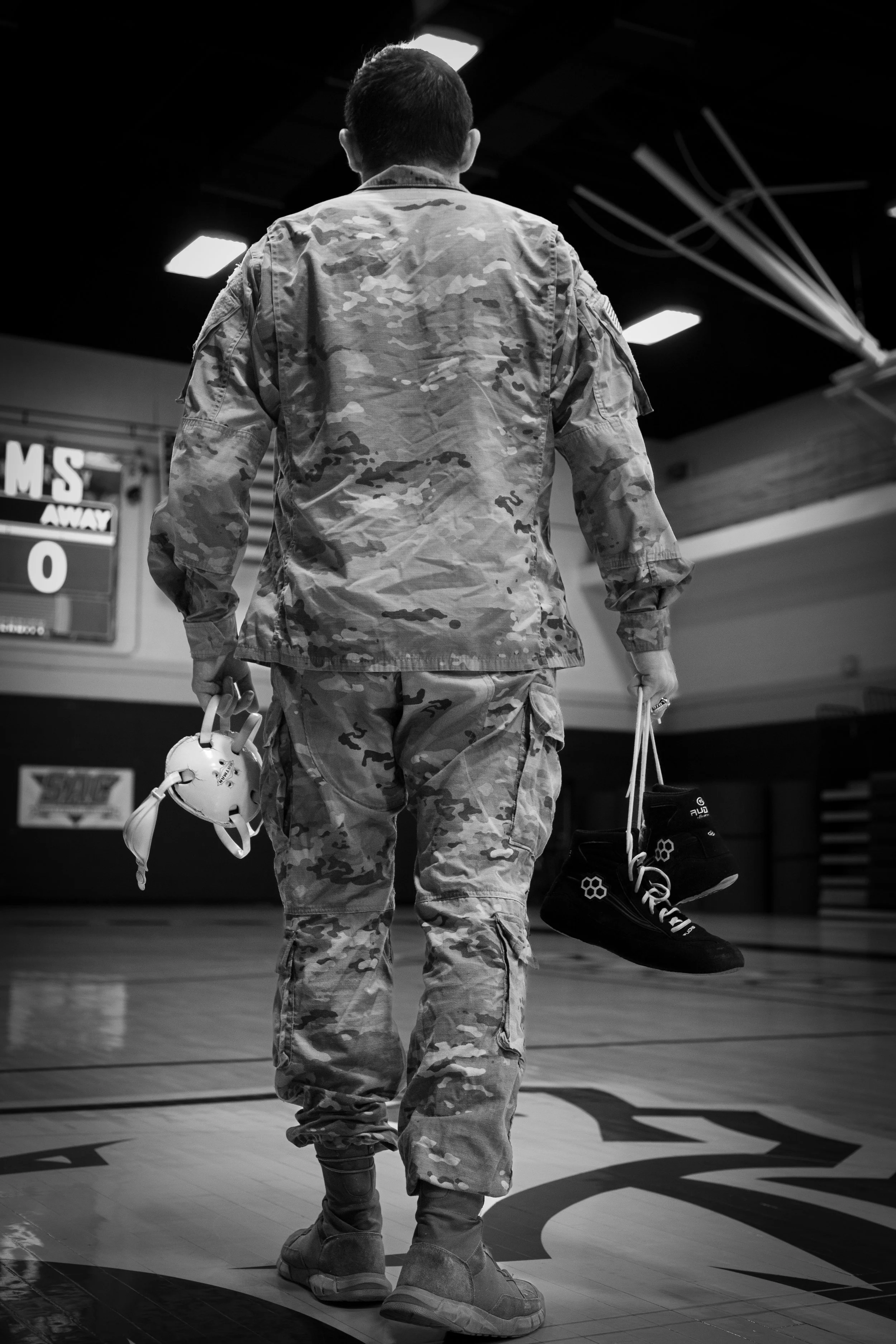 A soldier in camouflage uniform walking on a gymnasium floor, carrying a helmet in one hand and a pair of shoes in the other, with a scoreboard in the background.