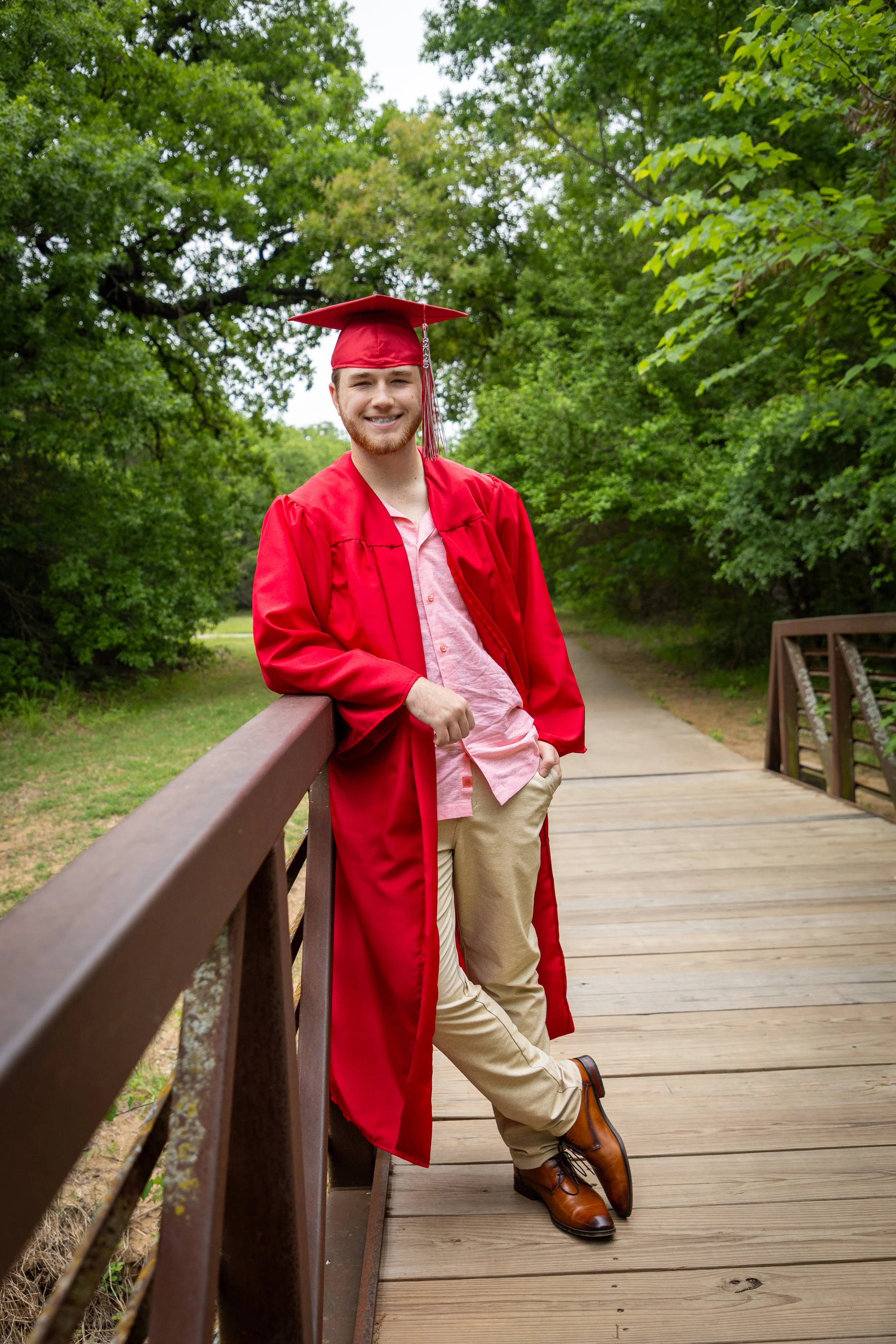Young man wearing a red graduation cap and gown, leaning on a railing on a wooden bridge, outdoors with green trees.