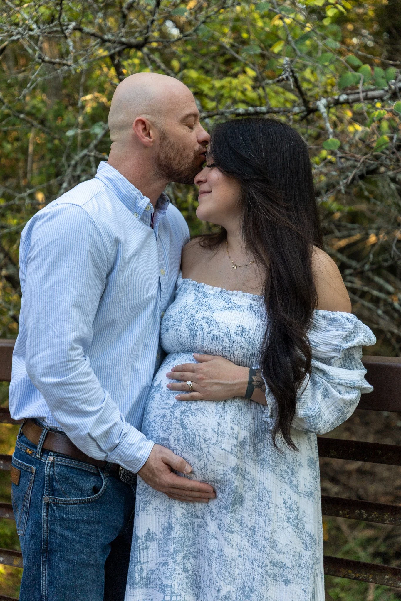 A man with a bald head and beard kisses a pregnant woman on her forehead outdoors.