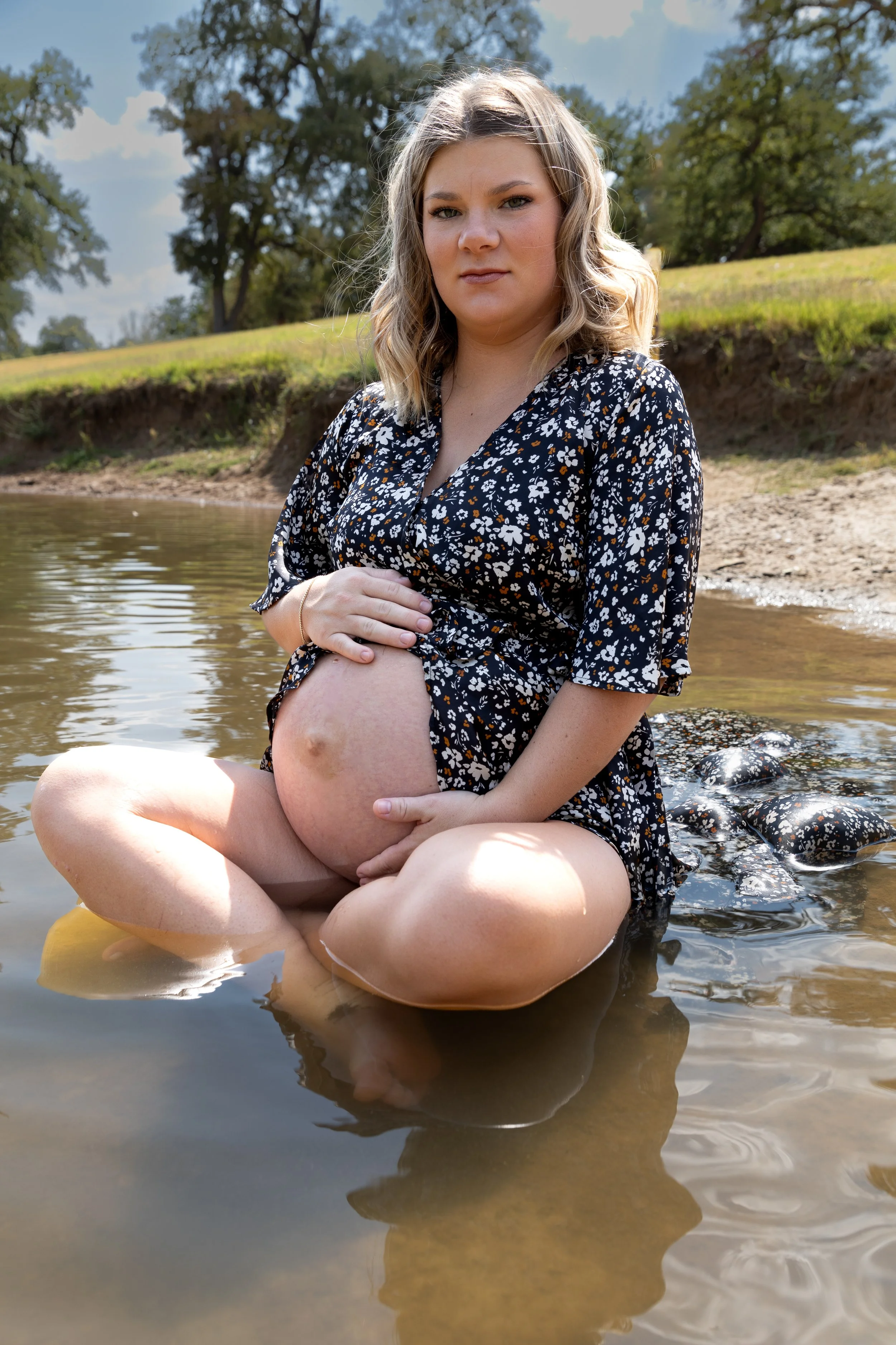 A pregnant woman with blonde wavy hair sitting in a shallow river during daytime, holding her belly with both hands, wearing a black floral dress, with a grassy hill and trees in the background.