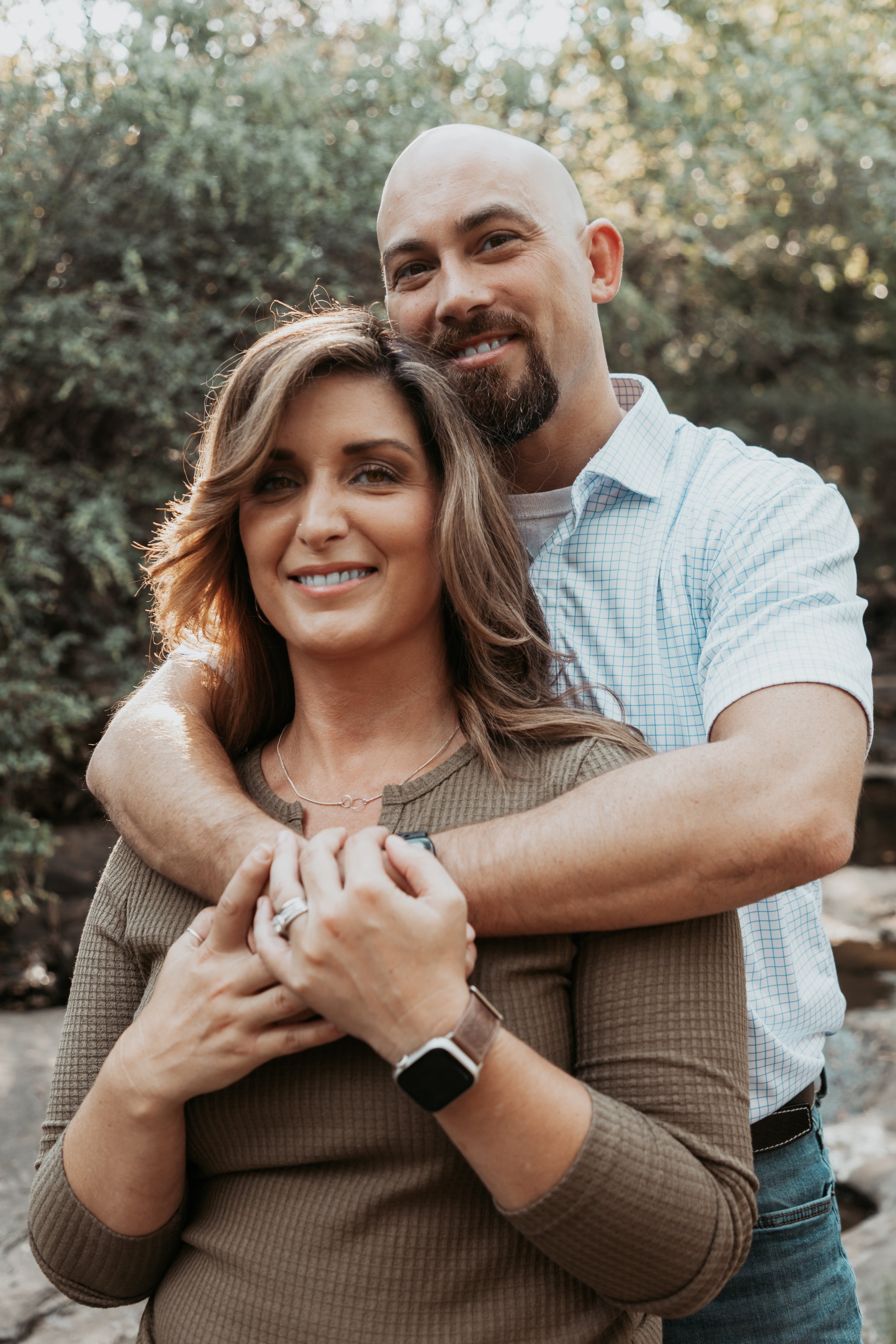 A smiling couple stands outside embracing each other, with a background of trees and sunlight filtering through the leaves.