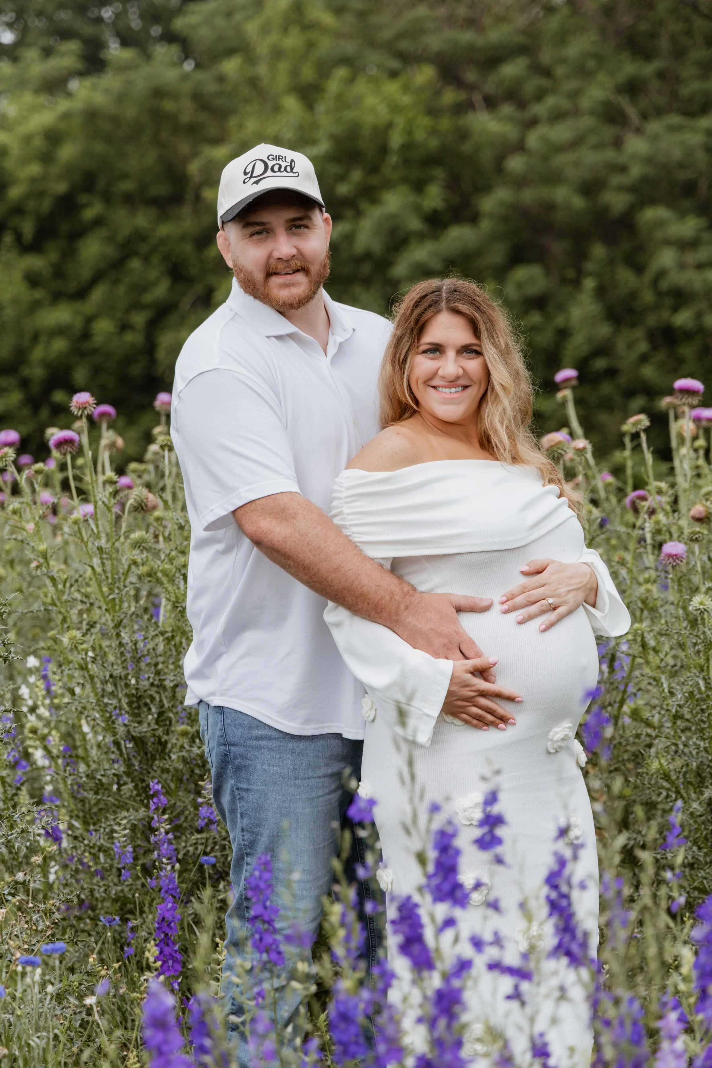 A pregnant woman with wavy blonde hair in a white dress, standing in a field of purple flowers, with a man in a white shirt and a cap that says 'Girl Dad' standing behind her, holding her belly.