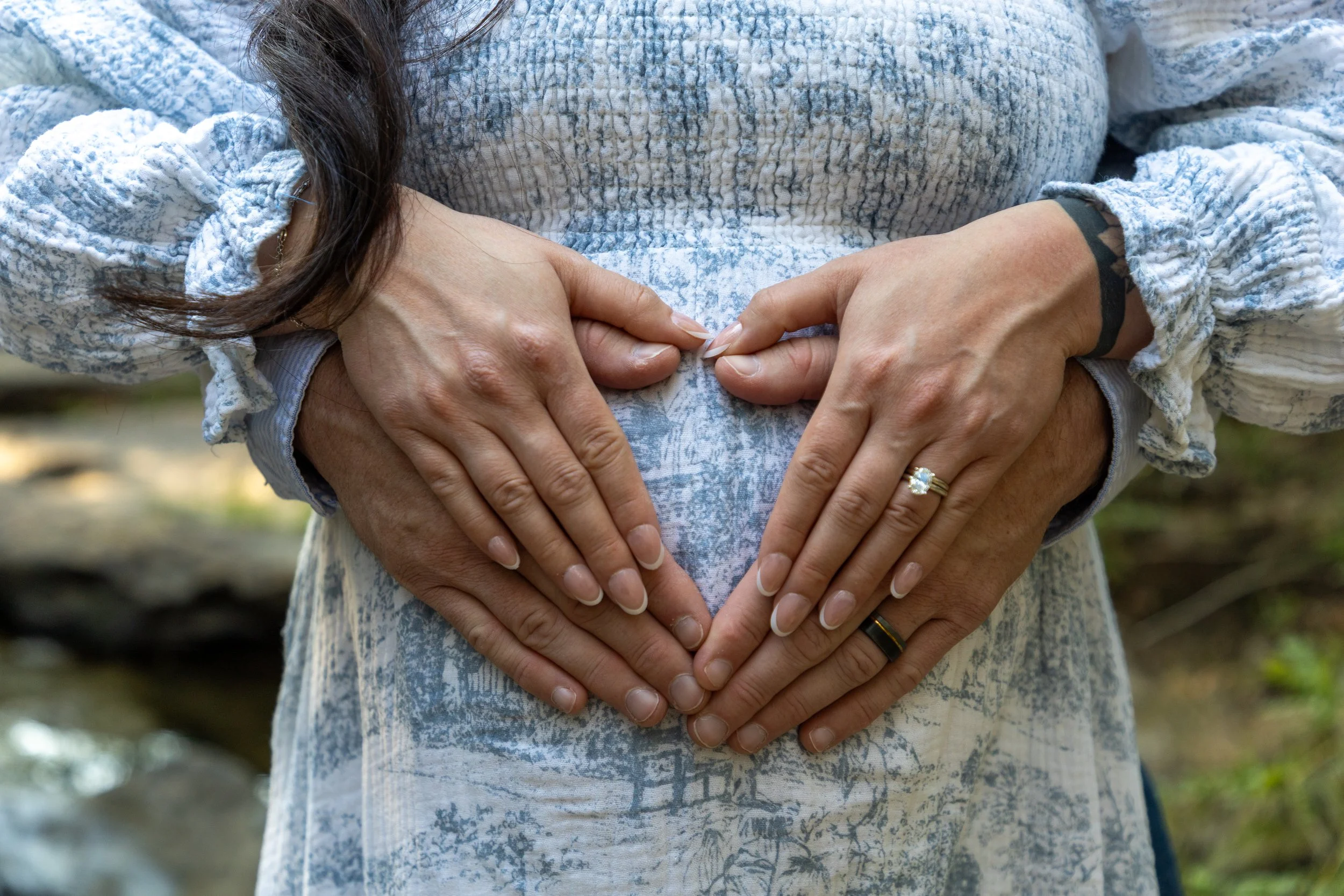 Two women forming a heart shape with their hands over their stomach, outdoors.