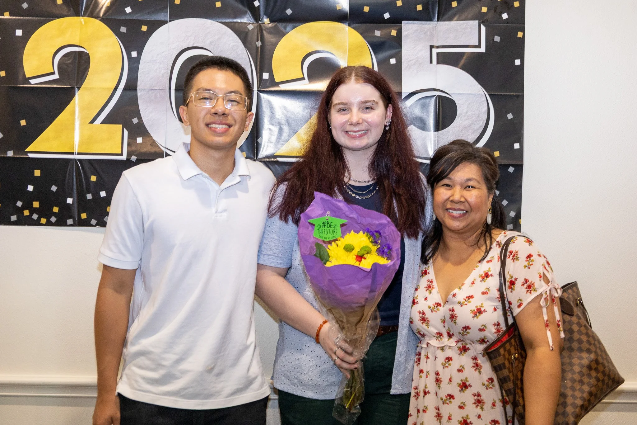Three women standing in front of a black and yellow '2023' celebration banner. The woman in the middle is holding a bouquet of yellow sunflowers wrapped in purple paper and is smiling. The woman on the left wears glasses and a white polo shirt, the w