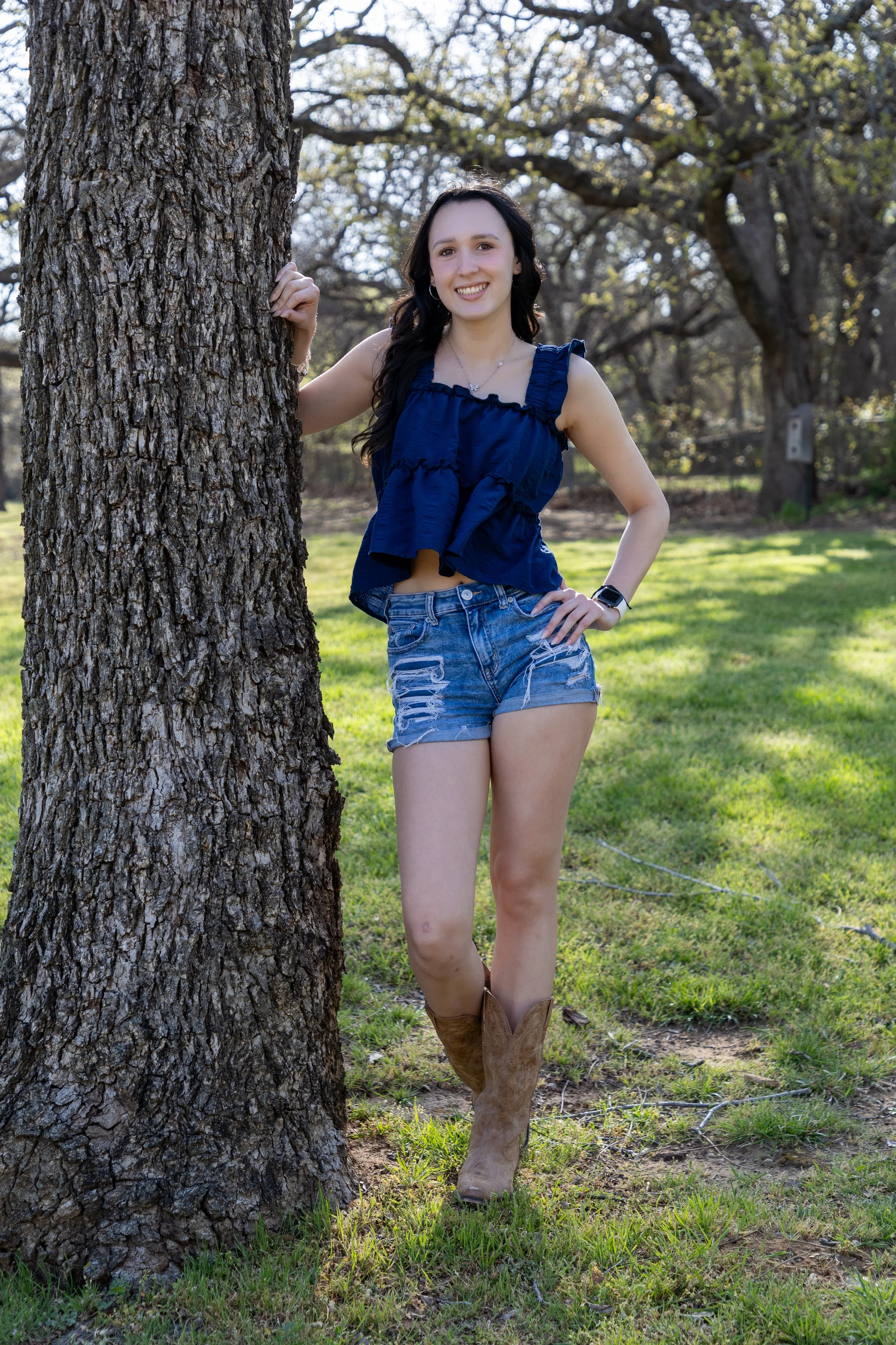A young woman smiling and standing outdoors next to a large tree, wearing a blue sleeveless top, denim shorts, cowboy boots, and a watch.