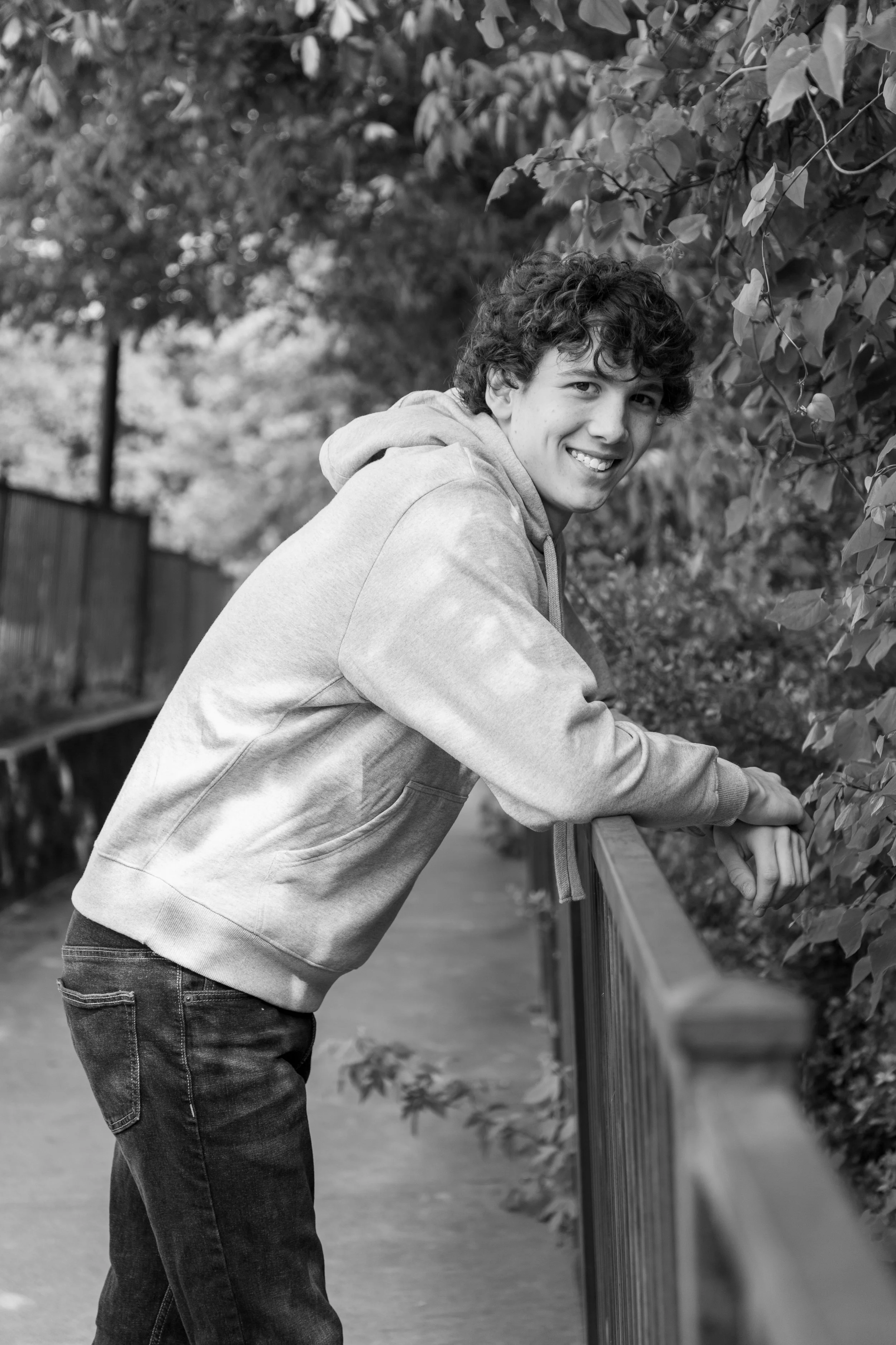 A young man with curly hair, smiling and leaning on a wooden fence, outdoors surrounded by trees.