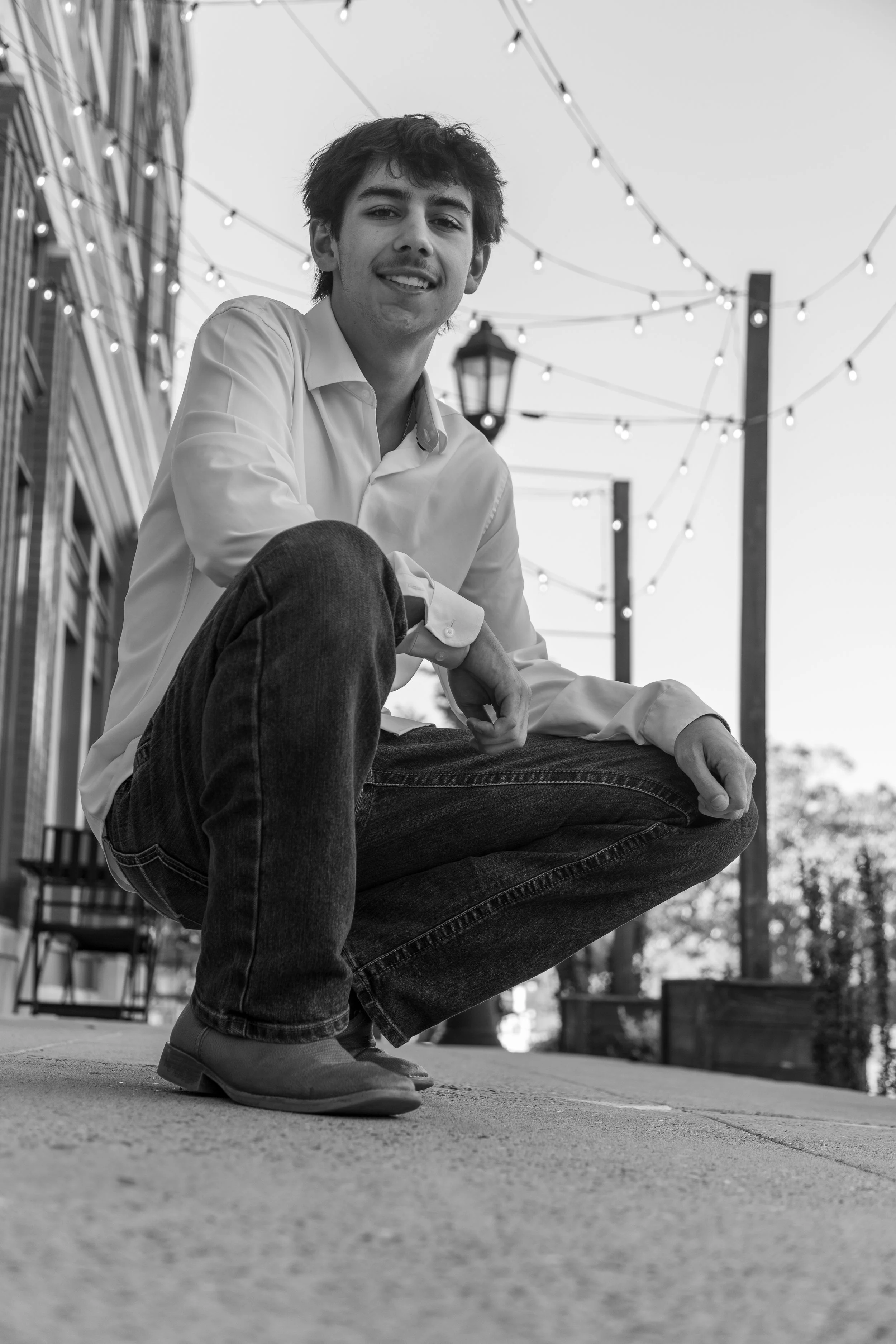 A young man crouching on a sidewalk, smiling, with string lights and a lamp post in the background.