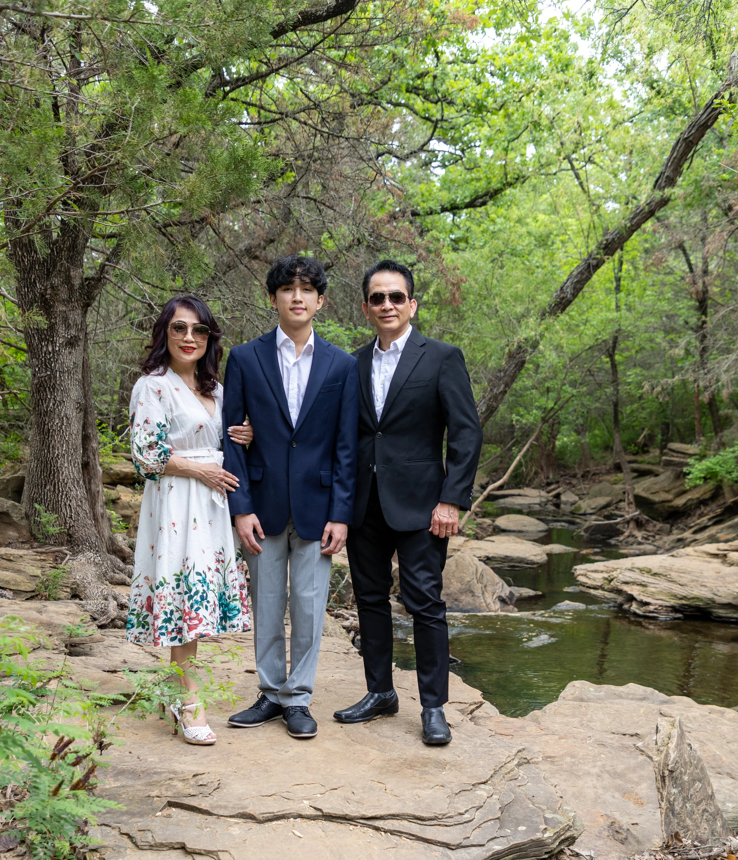 A family of three standing outdoors on rocks near a creek, surrounded by green trees. The woman is wearing sunglasses and a white floral dress, the son is in a navy suit and light gray pants, and the man is in a dark suit with sunglasses.