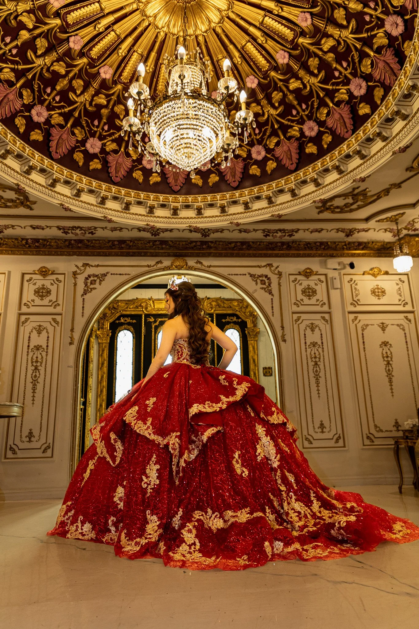A woman in a luxurious red and gold ball gown with an elaborate train, standing in an ornate, elegant ballroom with a chandelier and gold-accented decor.