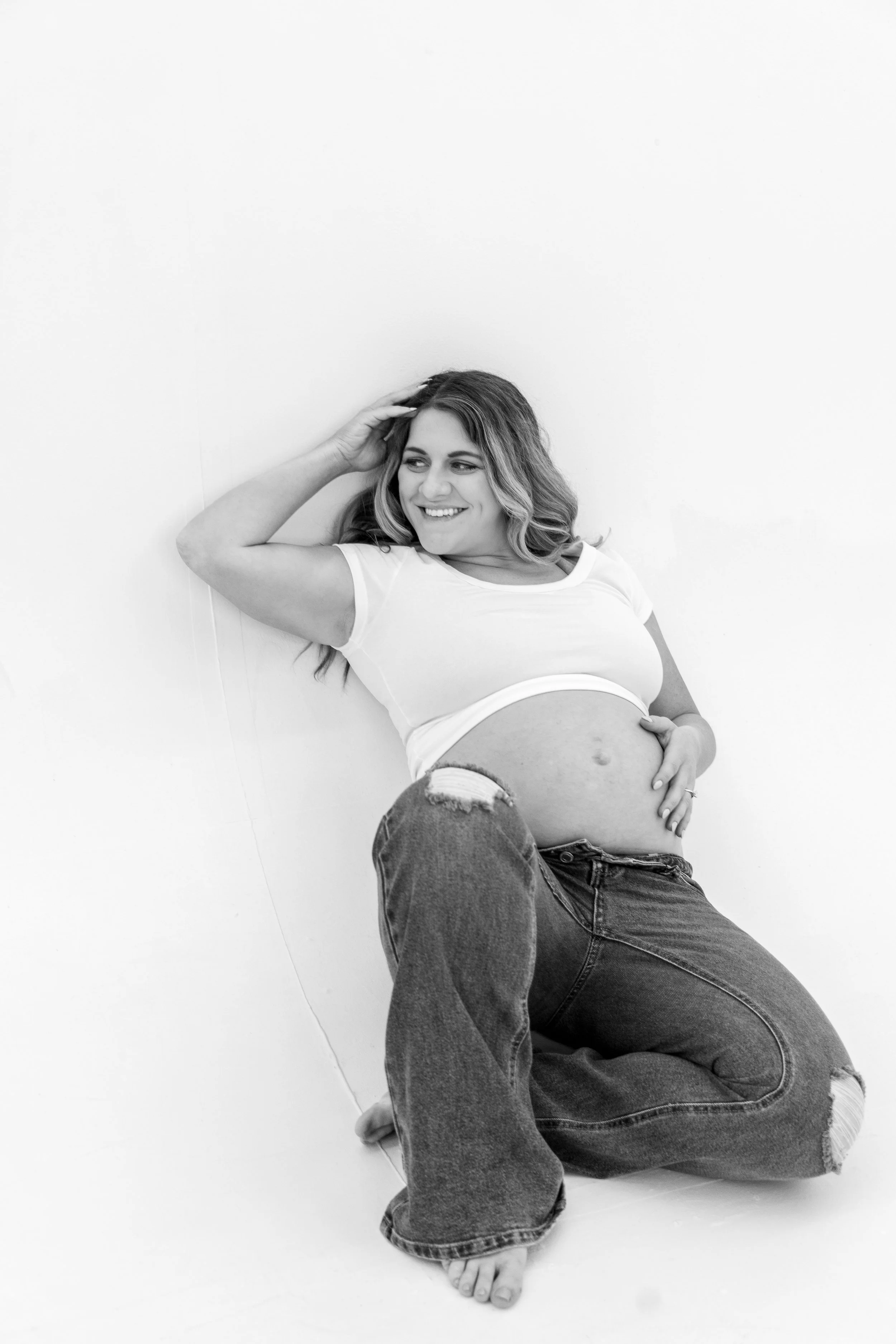 A woman with shoulder-length wavy hair, lying on the floor against a plain white background, smiling and wearing a white crop top and ripped jeans.