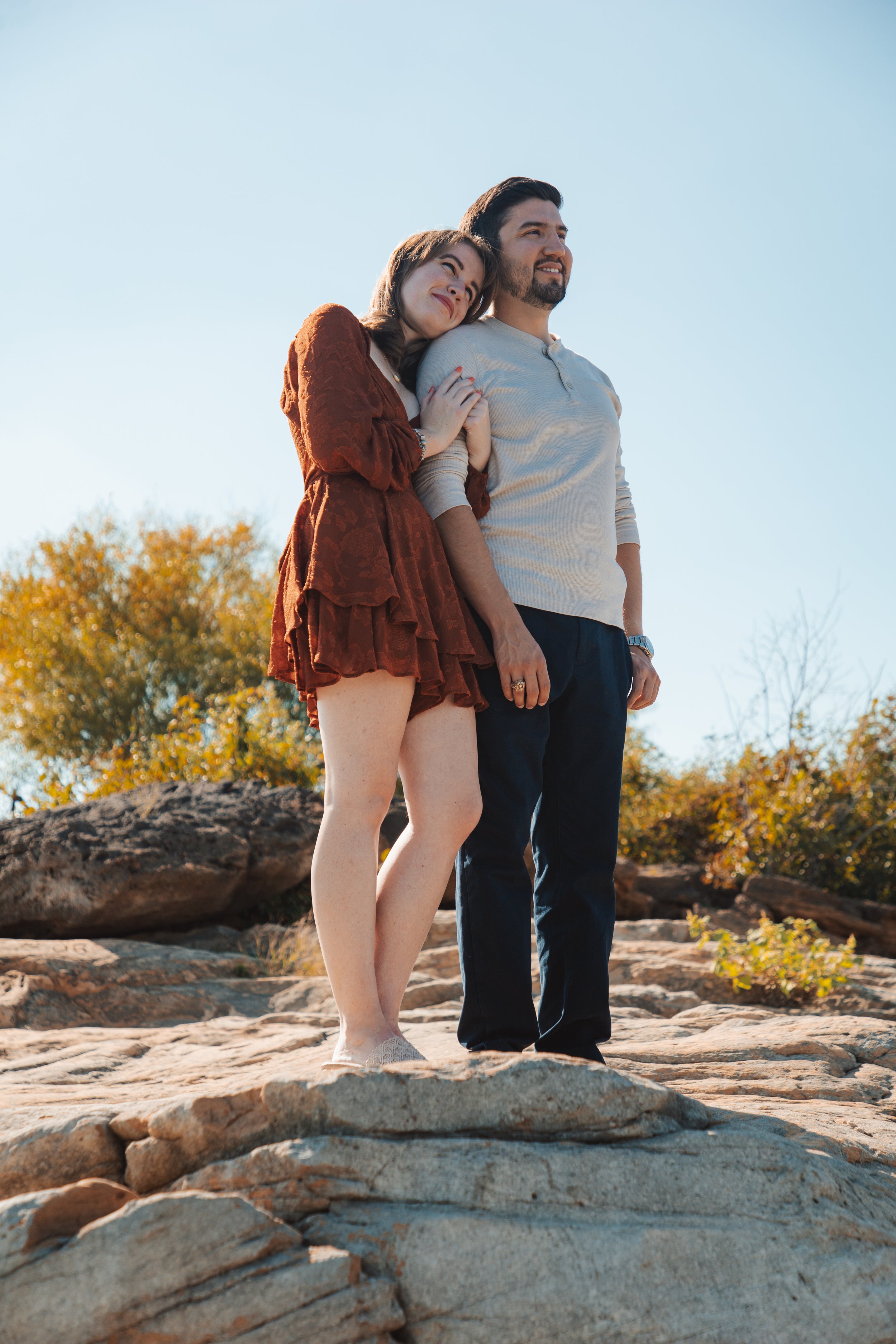 A couple standing on rocky terrain outdoors, with trees and a clear blue sky in the background. The woman is leaning her head on the man's shoulder, smiling and holding his arm, while the man looks off into the distance.