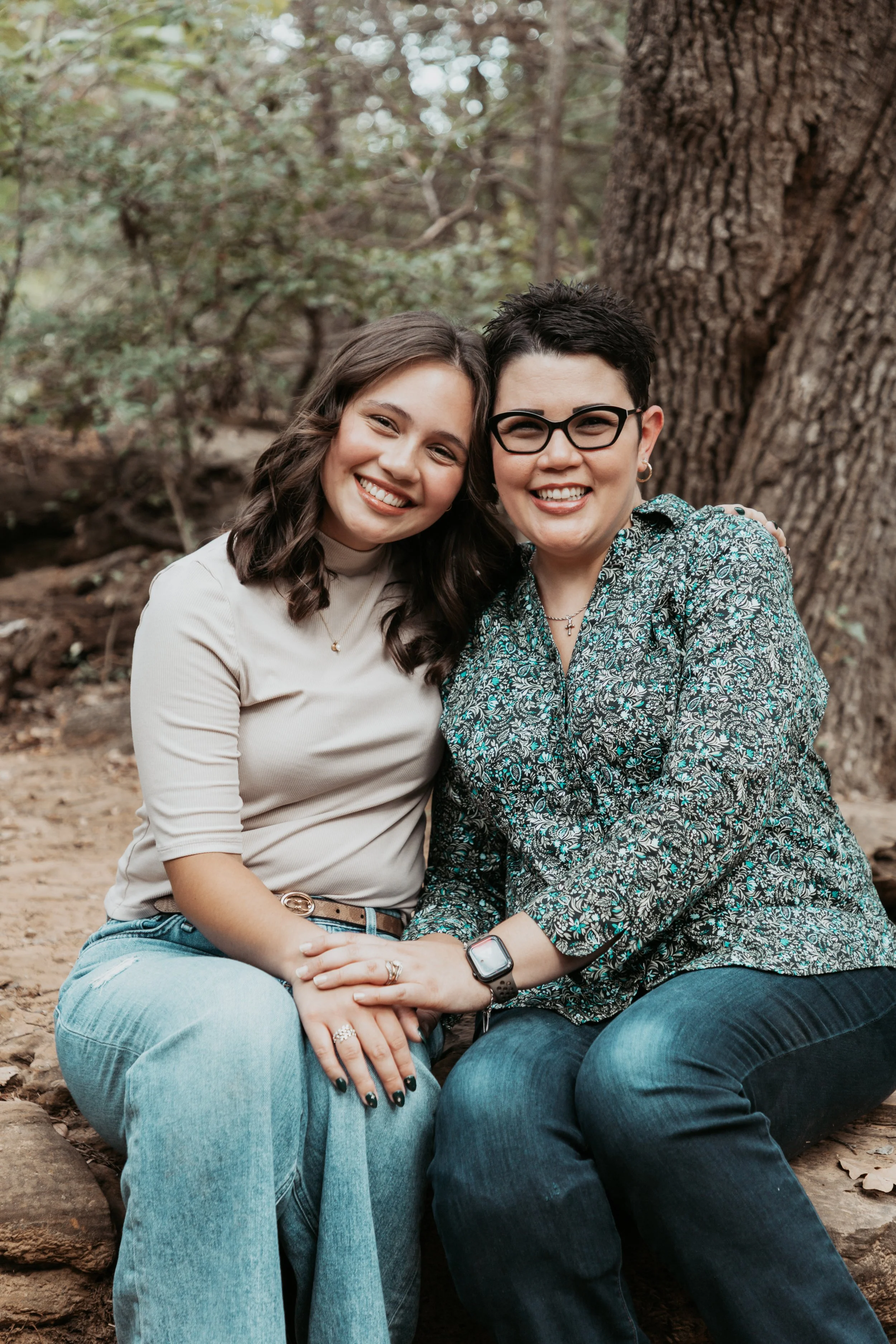 Two women sitting close together, smiling in a forested area with trees and rocks in the background.