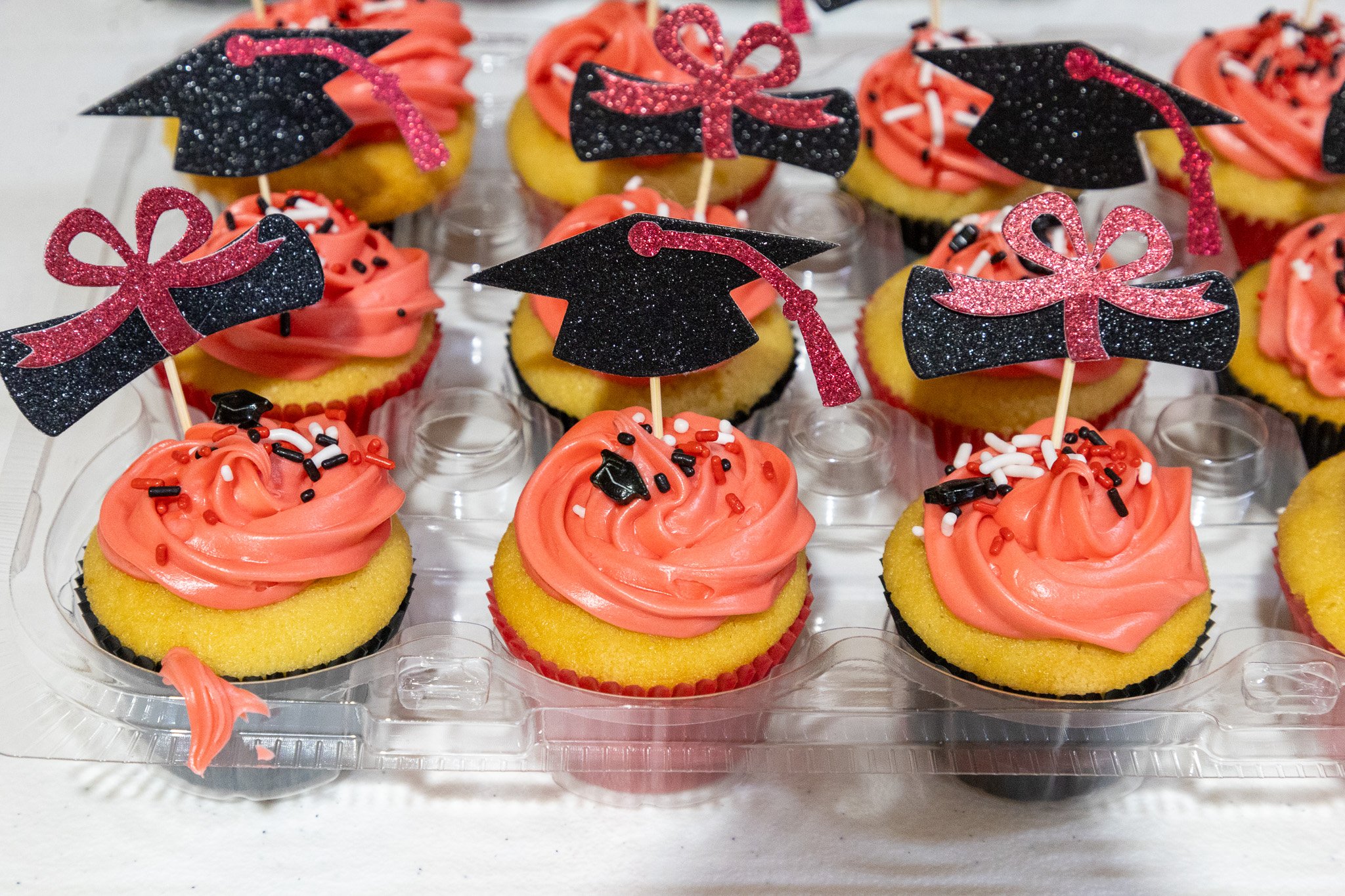 Cupcakes with pink frosting, black and pink glittery decoration toppers, some resembling graduation caps and diplomas, arranged in a clear plastic container.
