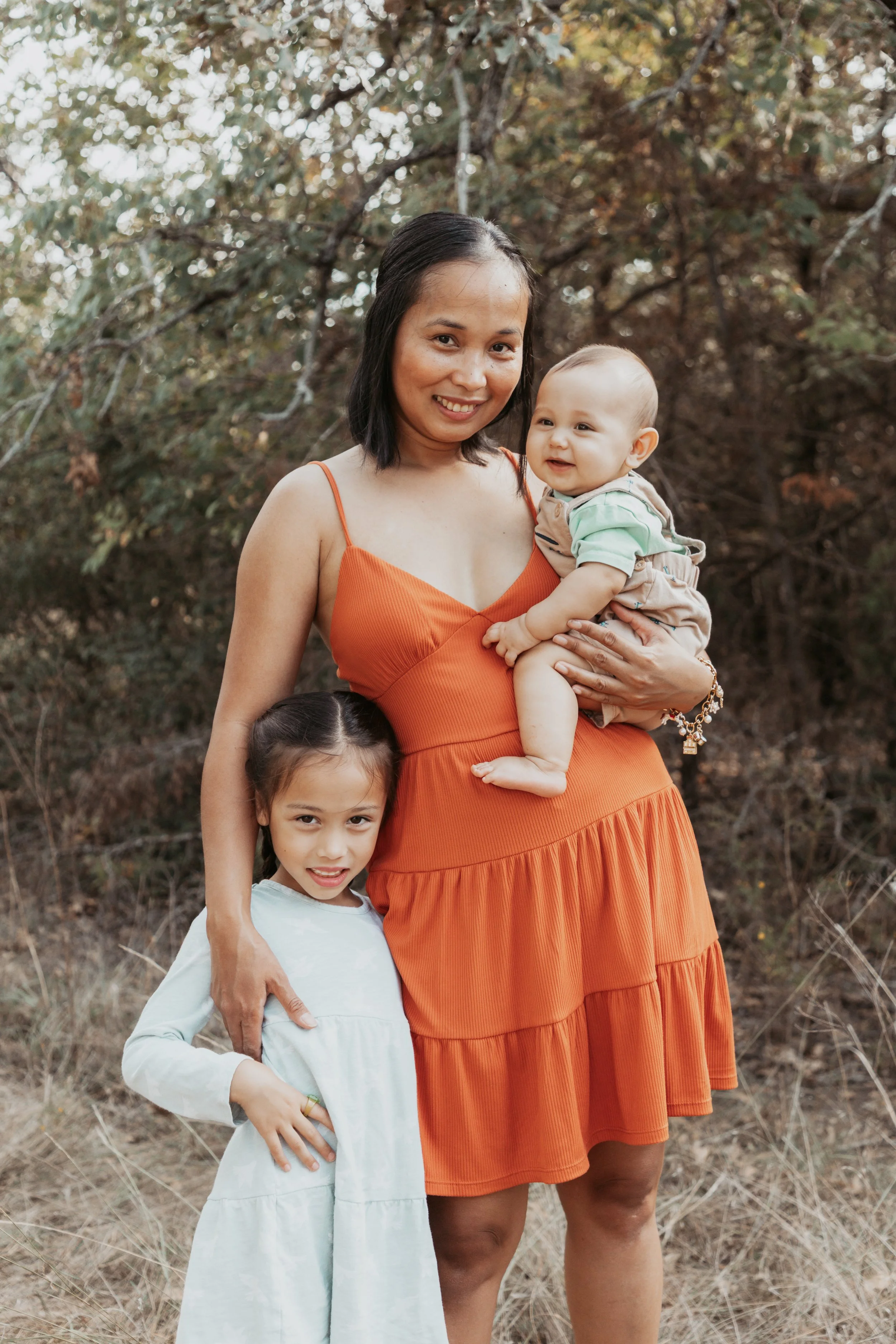 A woman in an orange dress holding a smiling baby, with a young girl standing beside her outdoors among trees.