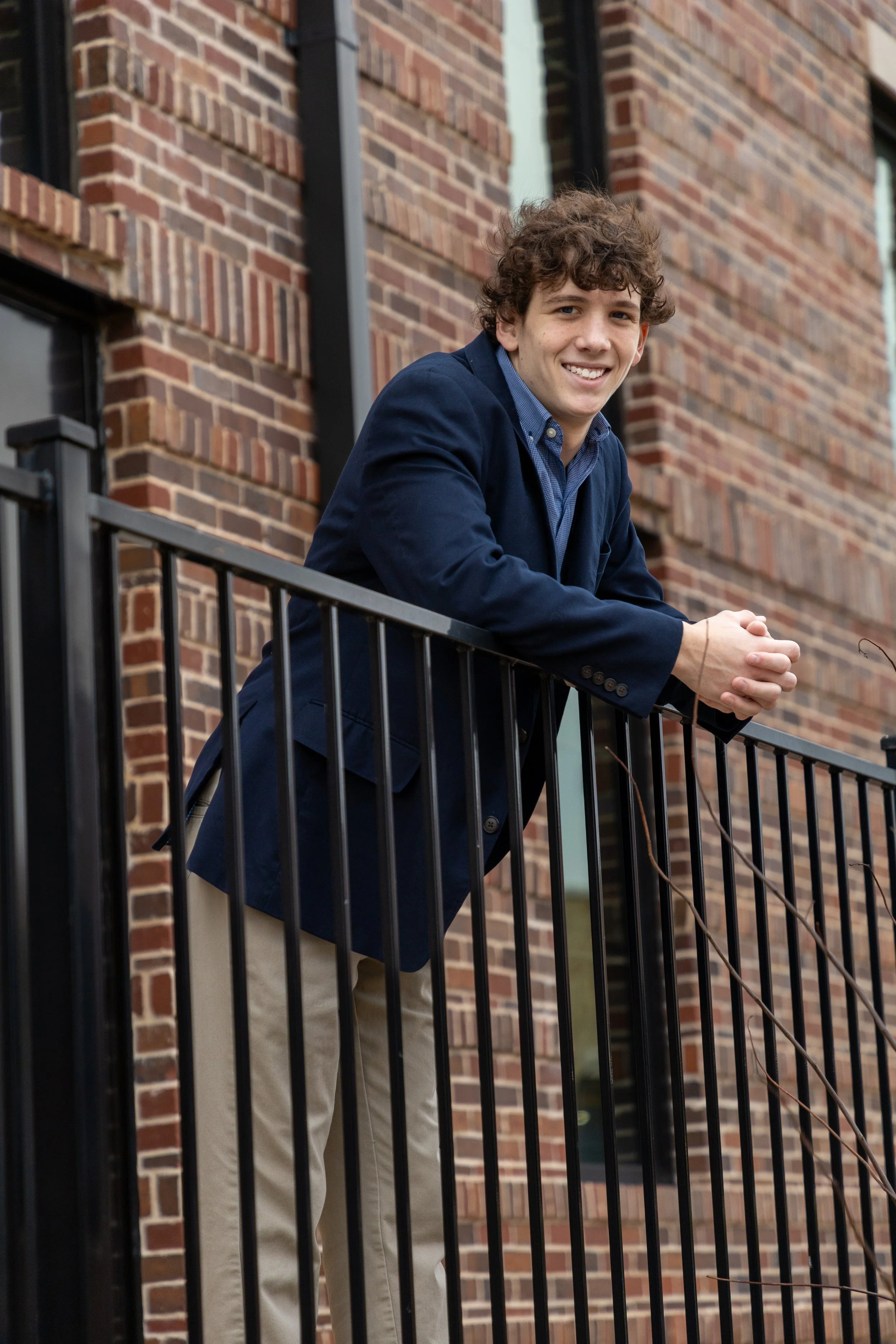 Young man with curly brown hair and a bright smile, leaning on a black metal railing outside a red brick building, dressed in a navy blue blazer and beige pants.