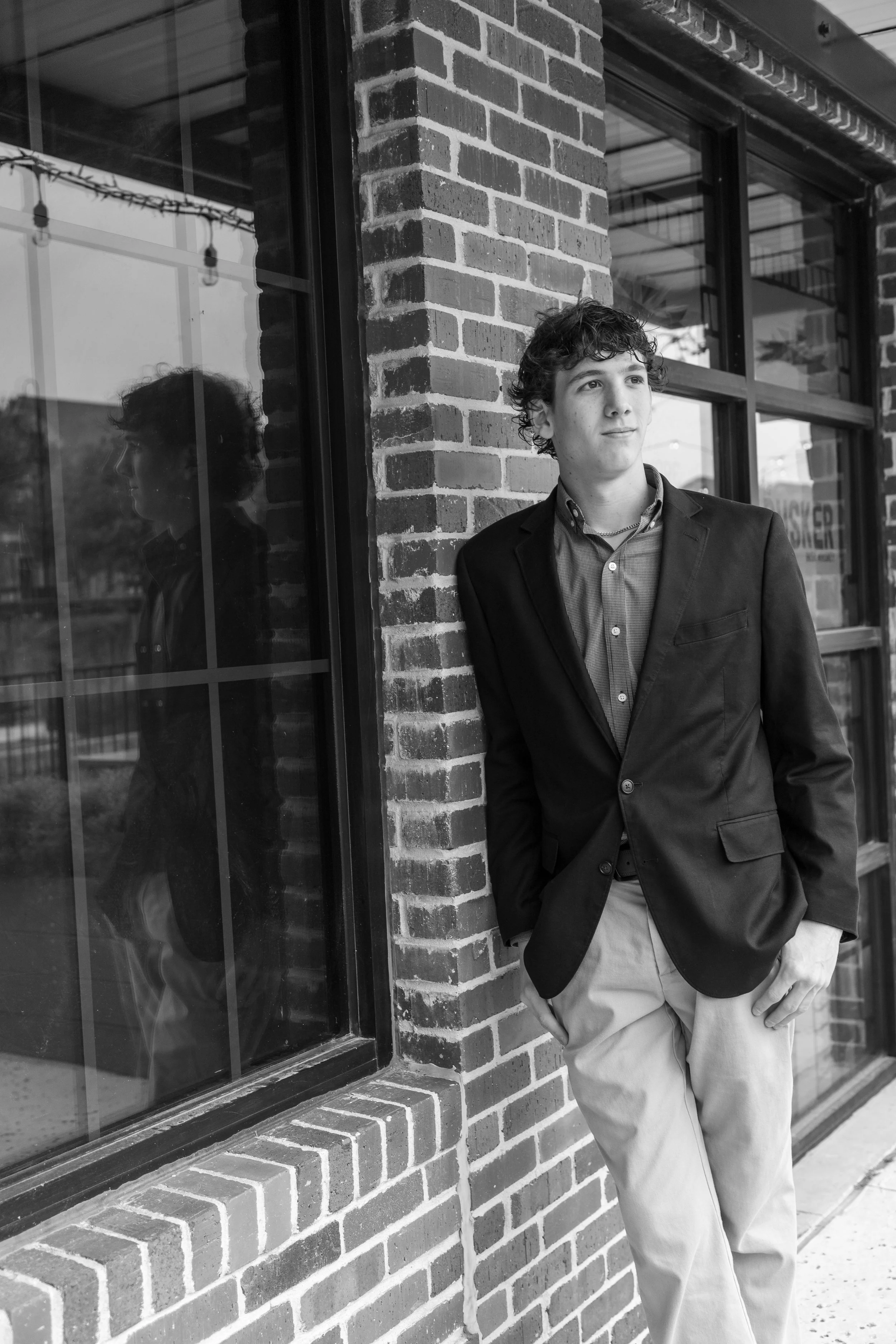 A young man in a blazer and khakis leans against a brick wall outside a building with large windows, reflecting his profile.