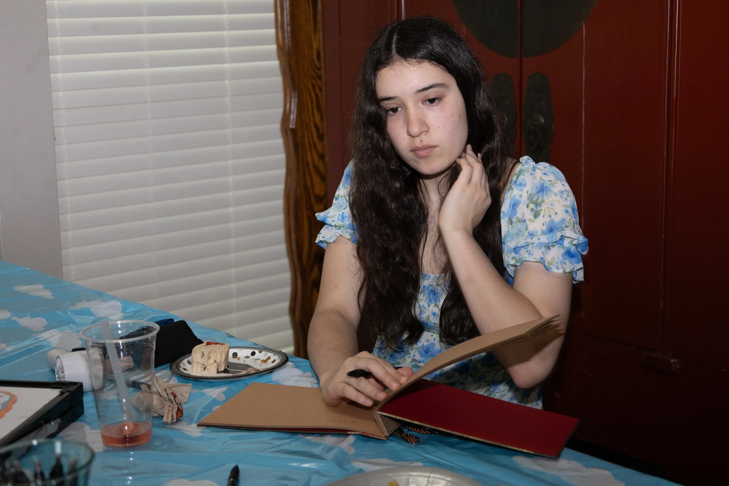 A young woman with long dark hair and light skin, wearing a blue floral dress, sitting at a table with a blue tablecloth, looking at a photo album and touching her neck with her left hand. On the table there is a plate with a piece of cake, a clear p