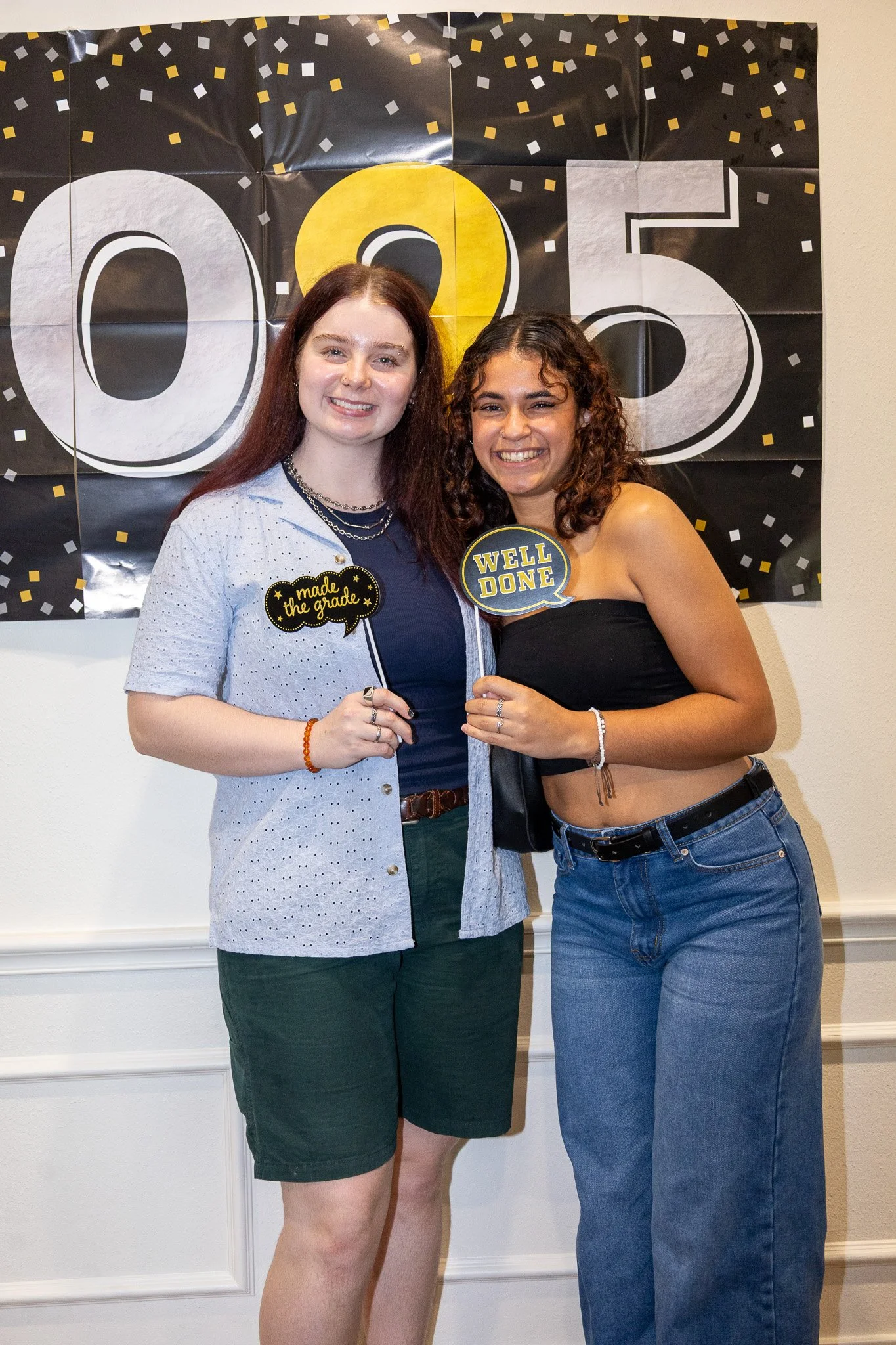 Two young women celebrating graduation, standing in front of a black and gold graduation banner with the number 2025. They are smiling, holding congratulatory signs that say 'made the grade' and 'well done,' dressed casually, with one in a black crop