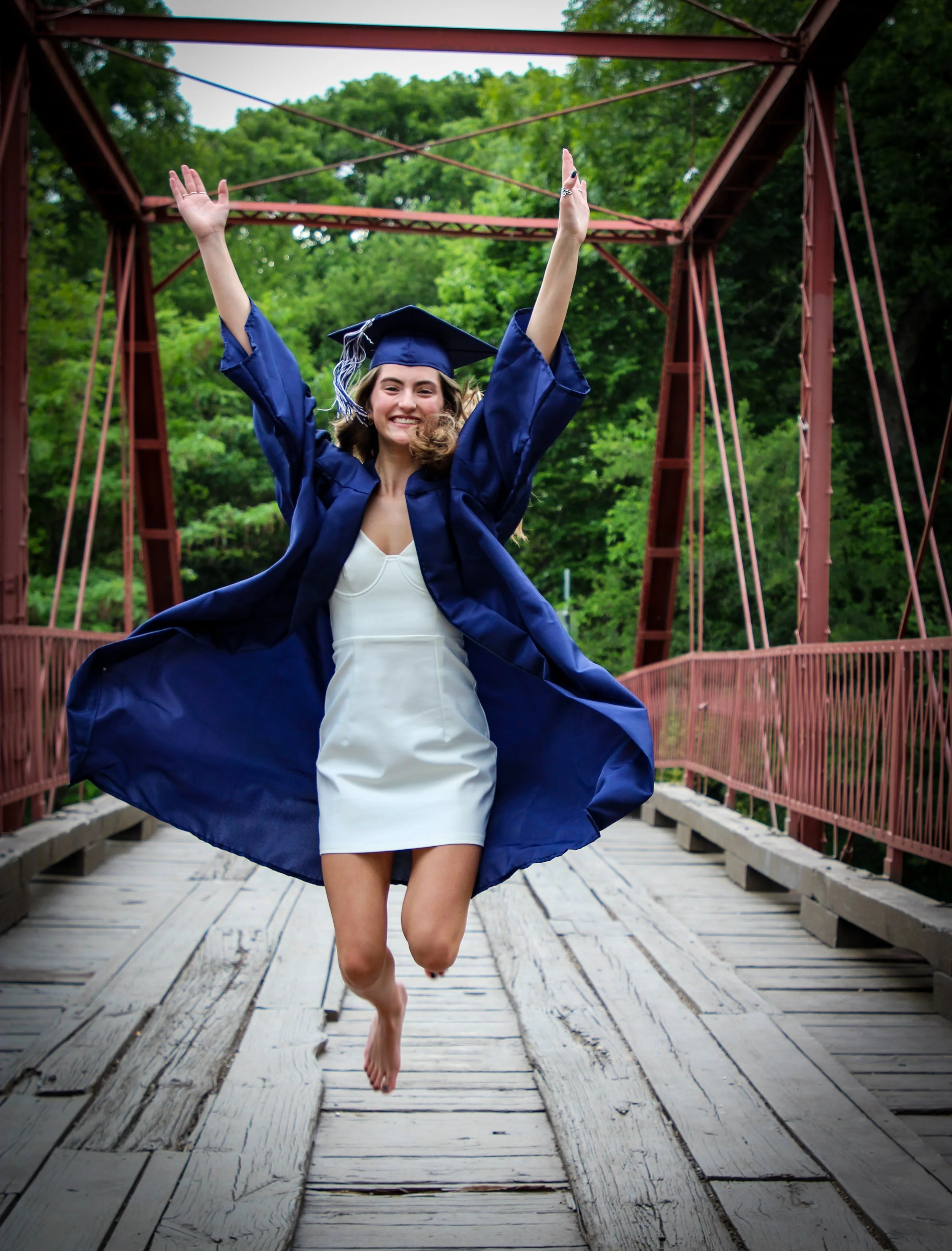 A young woman in a white dress and blue graduation gown and cap jumping on a wooden bridge, smiling with arms raised, surrounded by green trees.