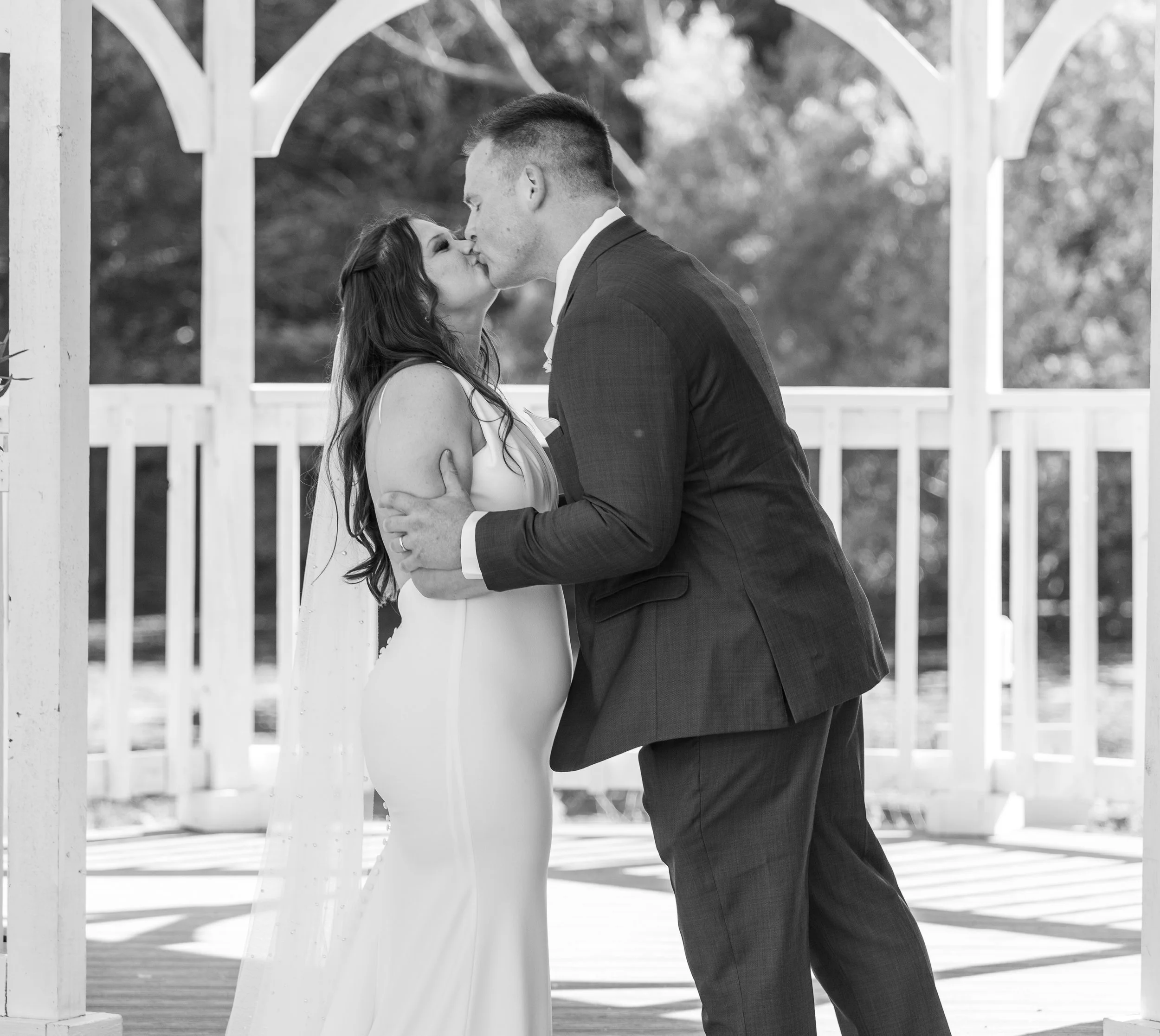 A black and white photo of a bride and groom kissing, holding each other, in an outdoor gazebo with a white fence and trees in the background.