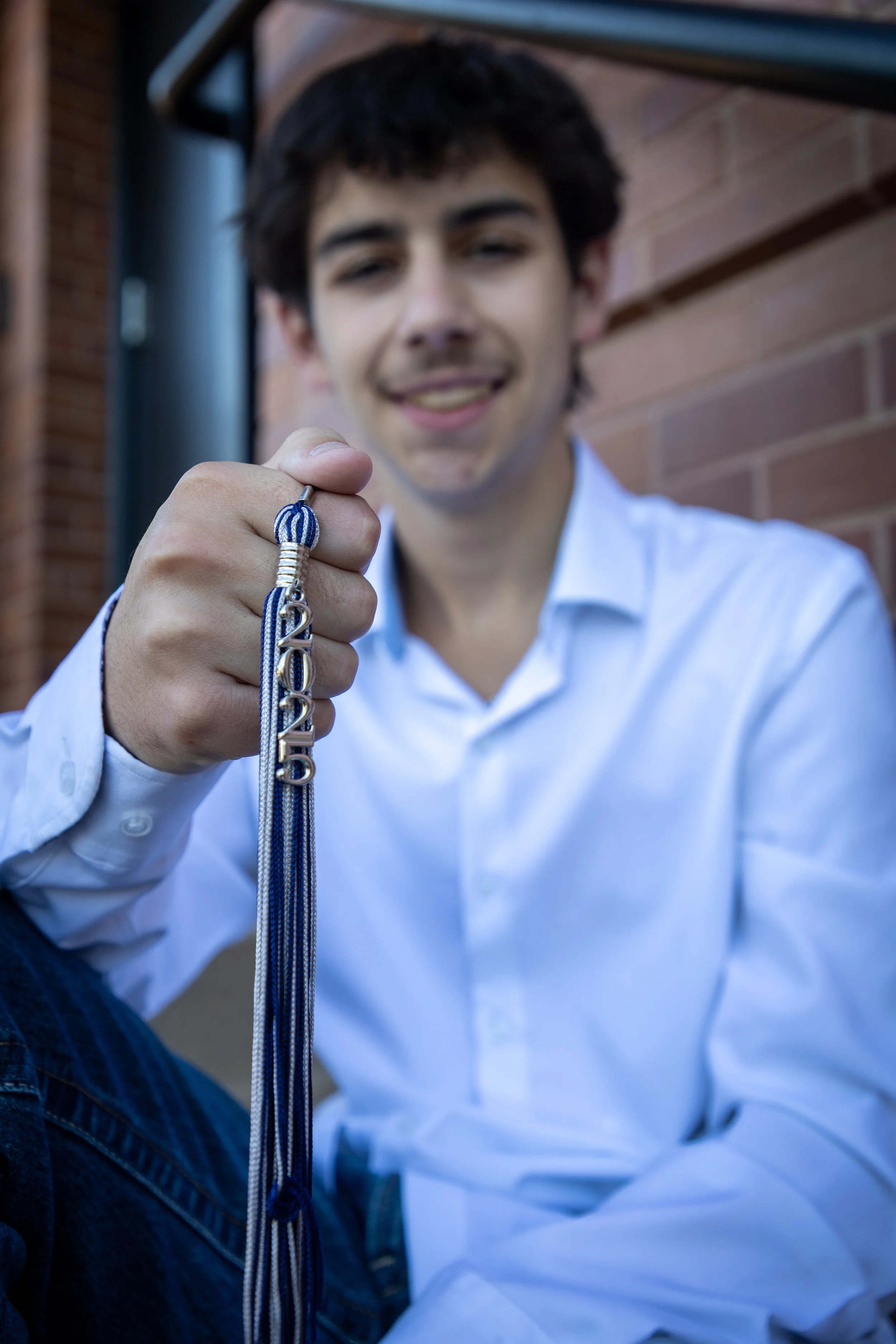 Young man in a white shirt holding a blue and white graduation cord with a silver "2023" charm, sitting outdoors with a brick wall background.