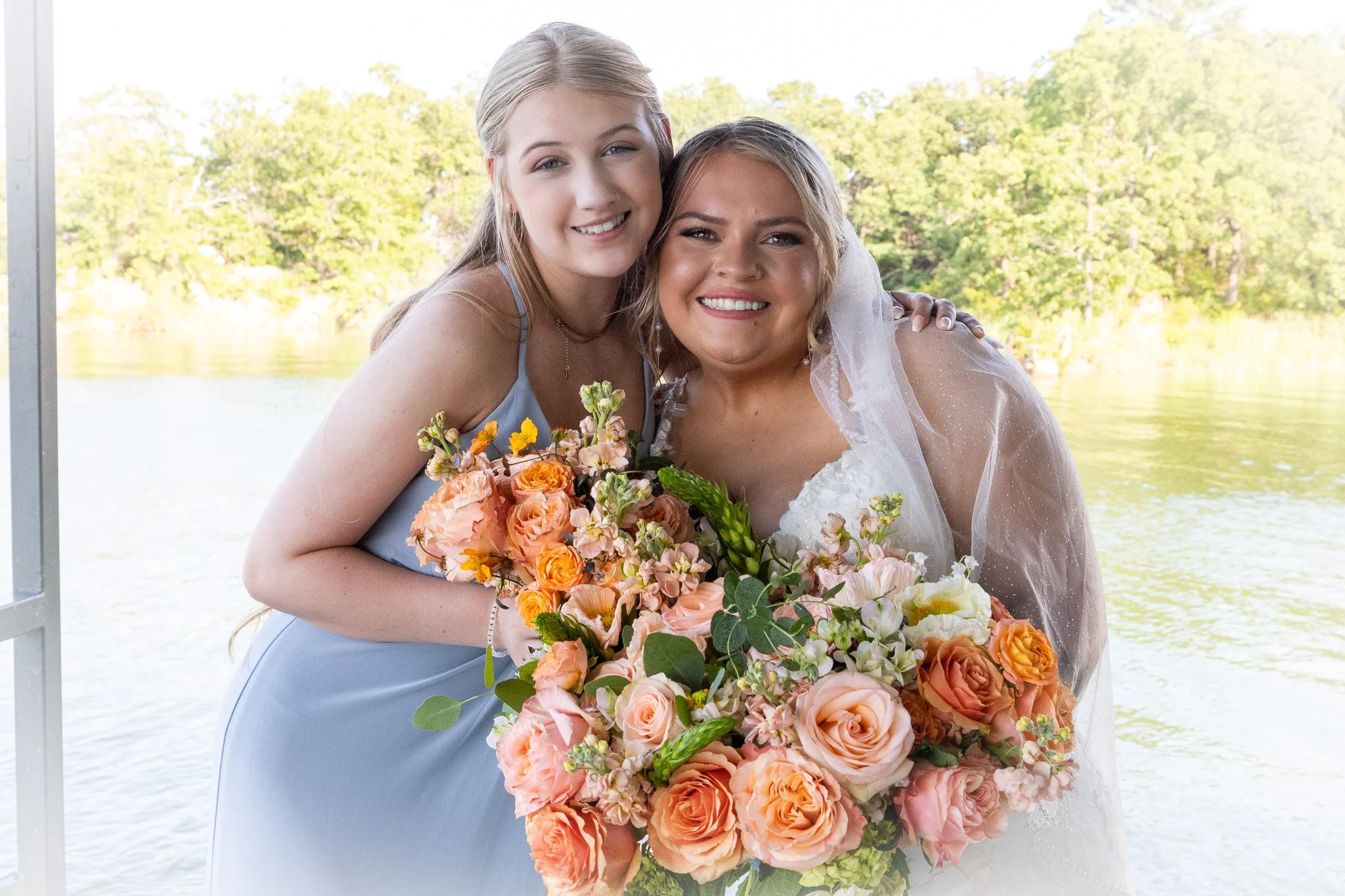 Two women smiling and hugging, one in a wedding dress with a veil and the other in a light blue dress, holding a large bouquet of pink, peach, and orange flowers, standing outdoors near a body of water with trees in the background.