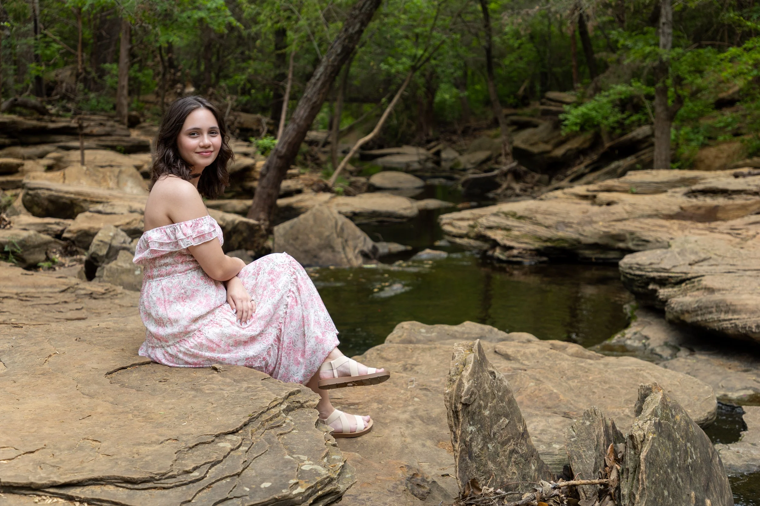 A woman in a pink floral off-the-shoulder dress sitting on a rock by a creek in a forest.