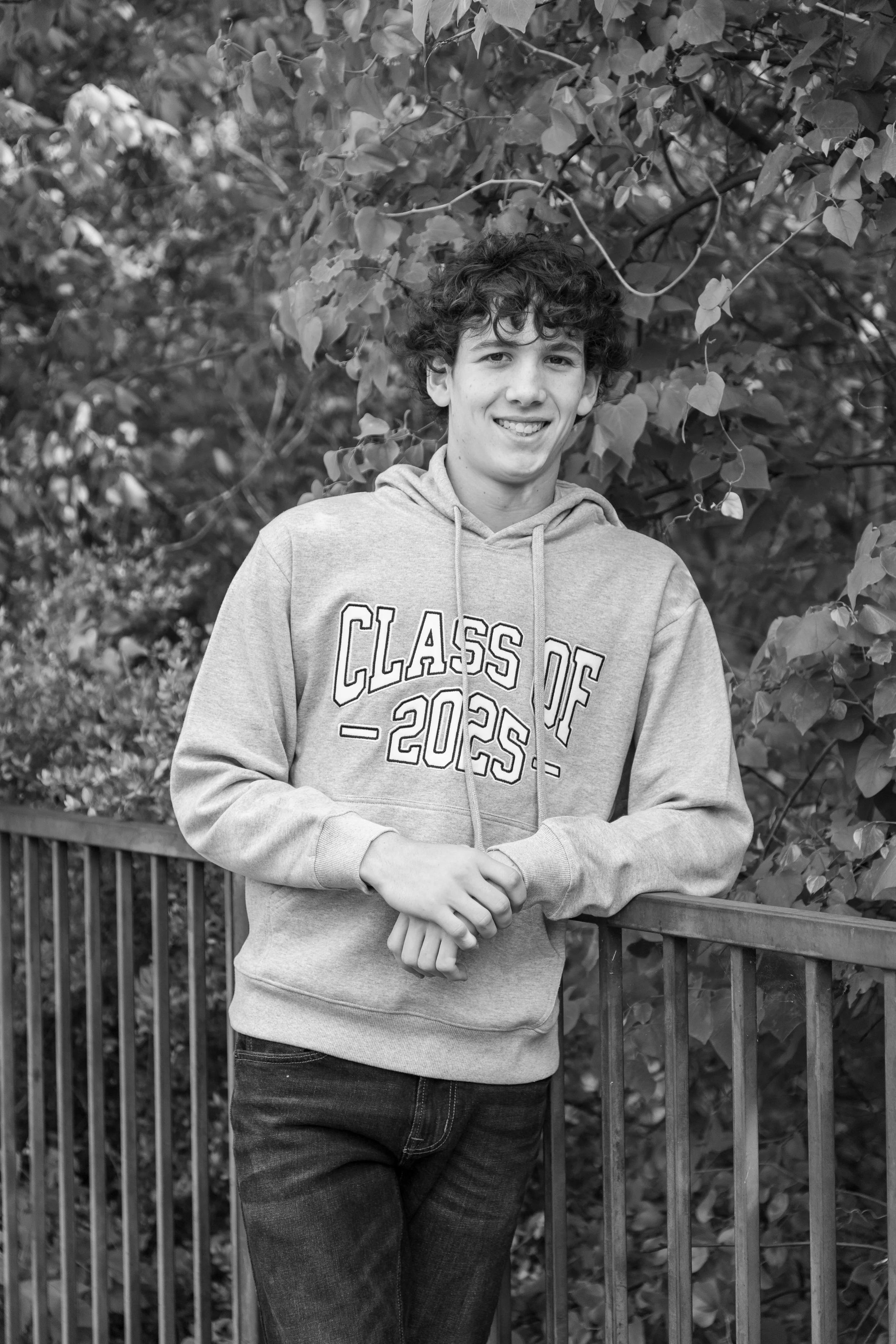 A young man with curly hair smiling, wearing a hooded sweatshirt with 'Class of 2023' written on it, standing outdoors beside a wooden railing with foliage in the background.