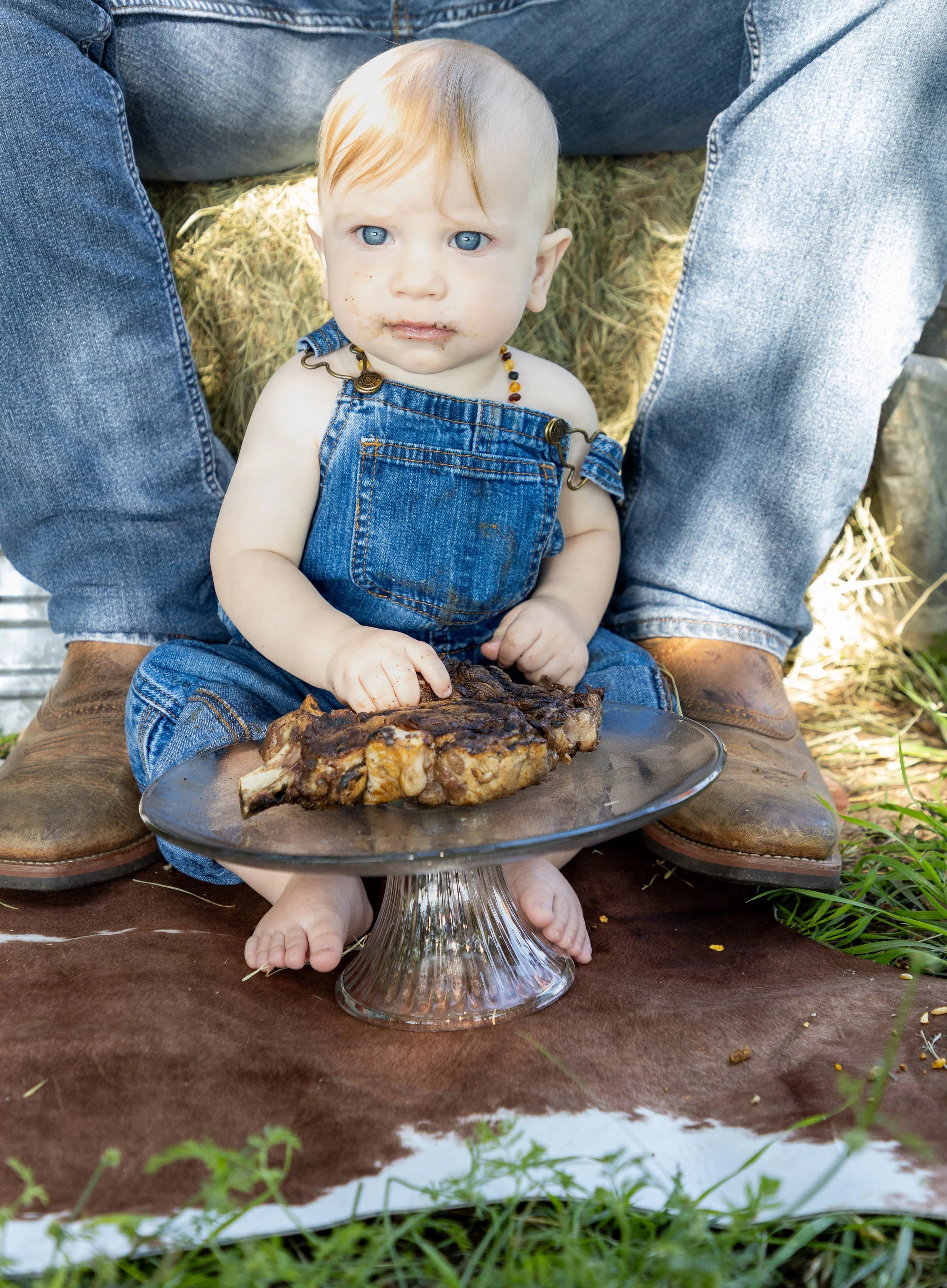 A baby with blue eyes and blonde hair, dressed in denim overalls, sitting on a brown cloth outdoors. The baby has food around their mouth and is touching a large cooked piece of meat on a glass cake stand. An adult, wearing blue jeans and brown boots