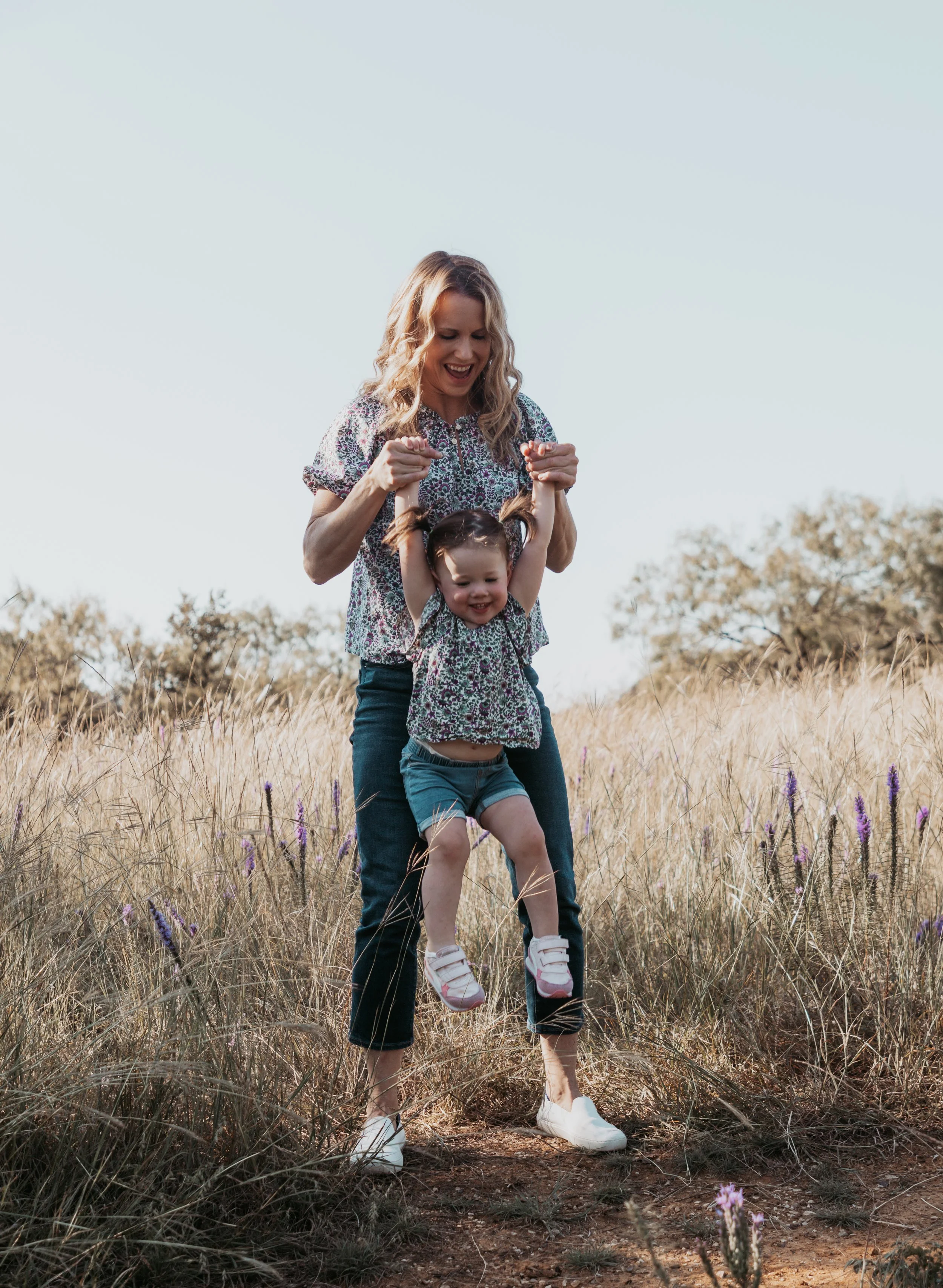 A woman and a young girl are in a field of tall grass and purple flowers. The woman is holding the girl up by her hands and they are both smiling, enjoying the moment.