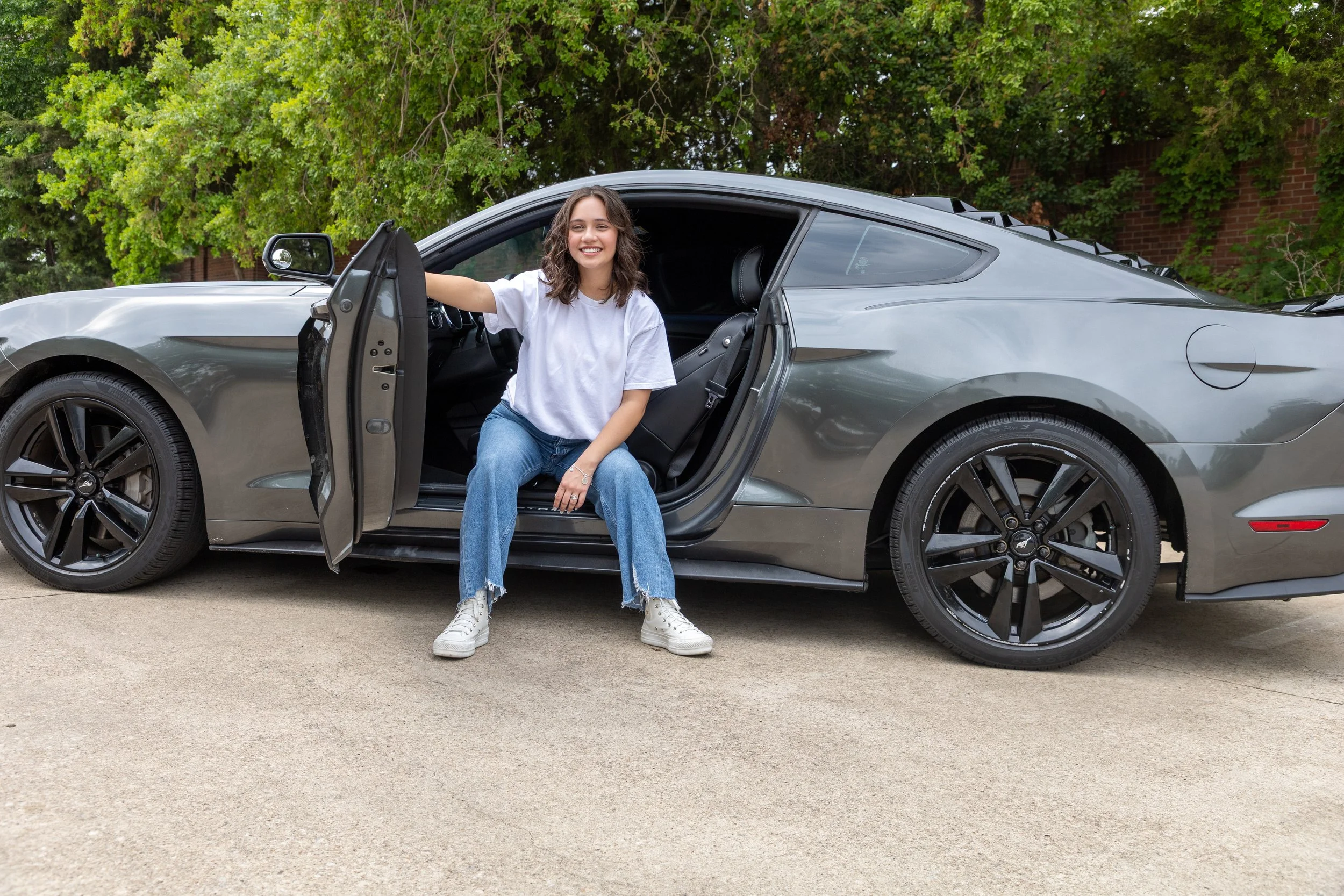 A young woman with shoulder-length brown hair, wearing a white t-shirt and blue jeans, sitting on the edge of an open car door of a sleek gray sports car, smiling happily. The setting is outdoors on a paved surface with green trees and part of a bric
