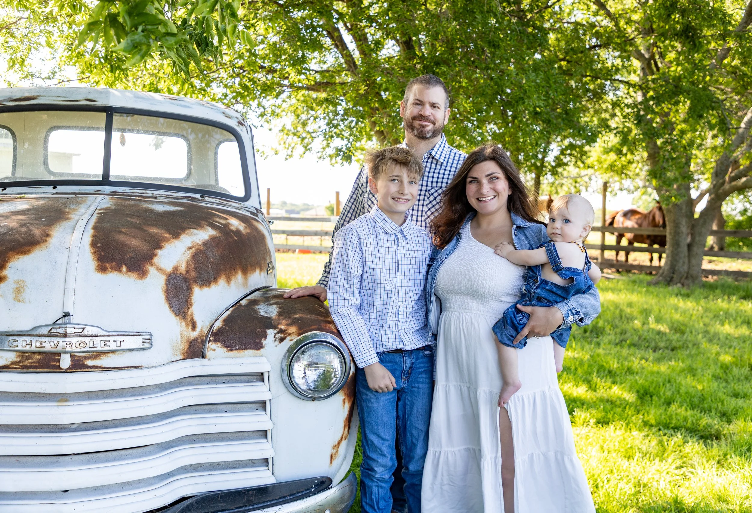 A family of four standing outdoors next to an old, rusty Chevrolet truck under a large tree, with horses visible in the background on a farm.