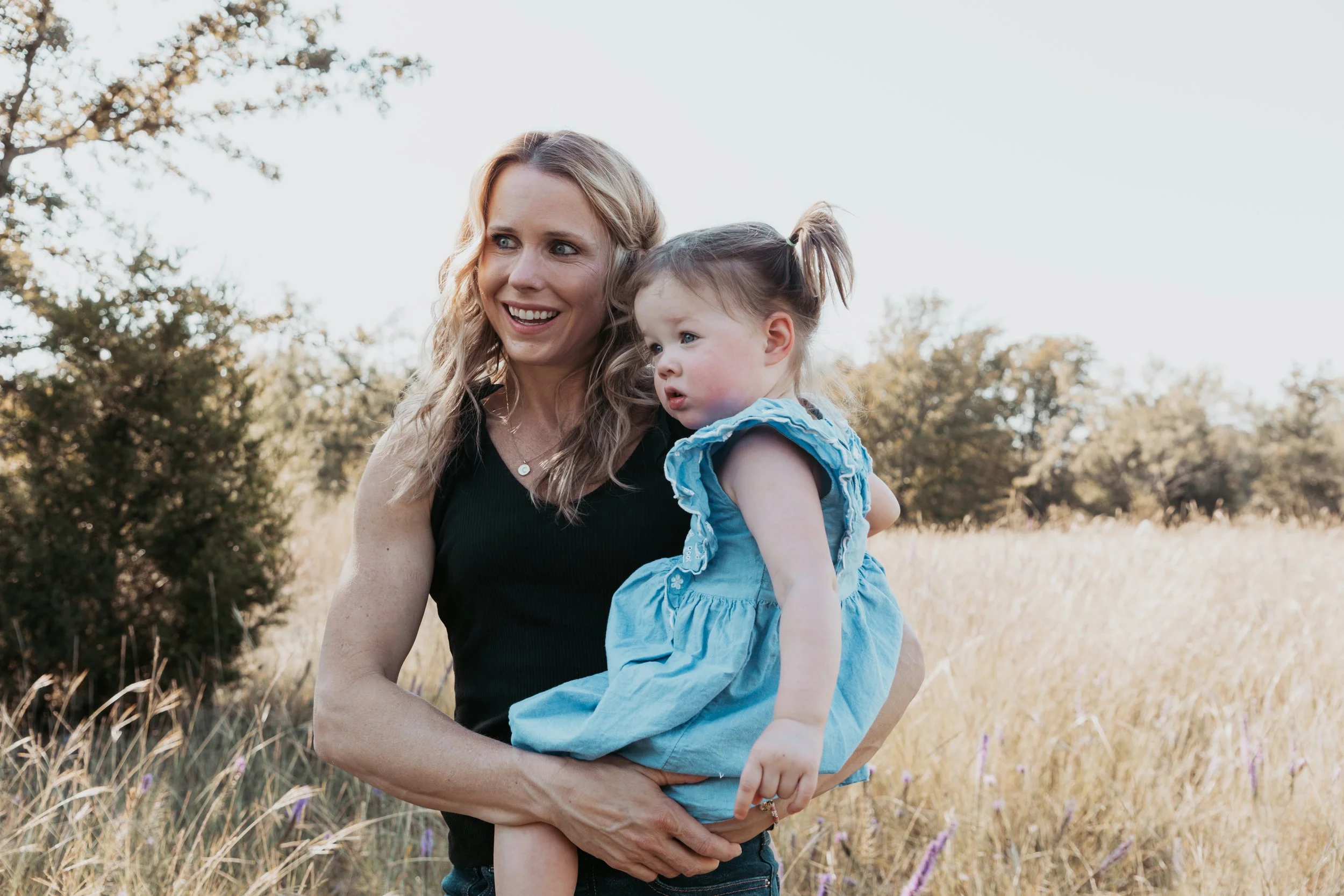 A woman holding a young girl in an outdoor field with tall grass and trees in the background.
