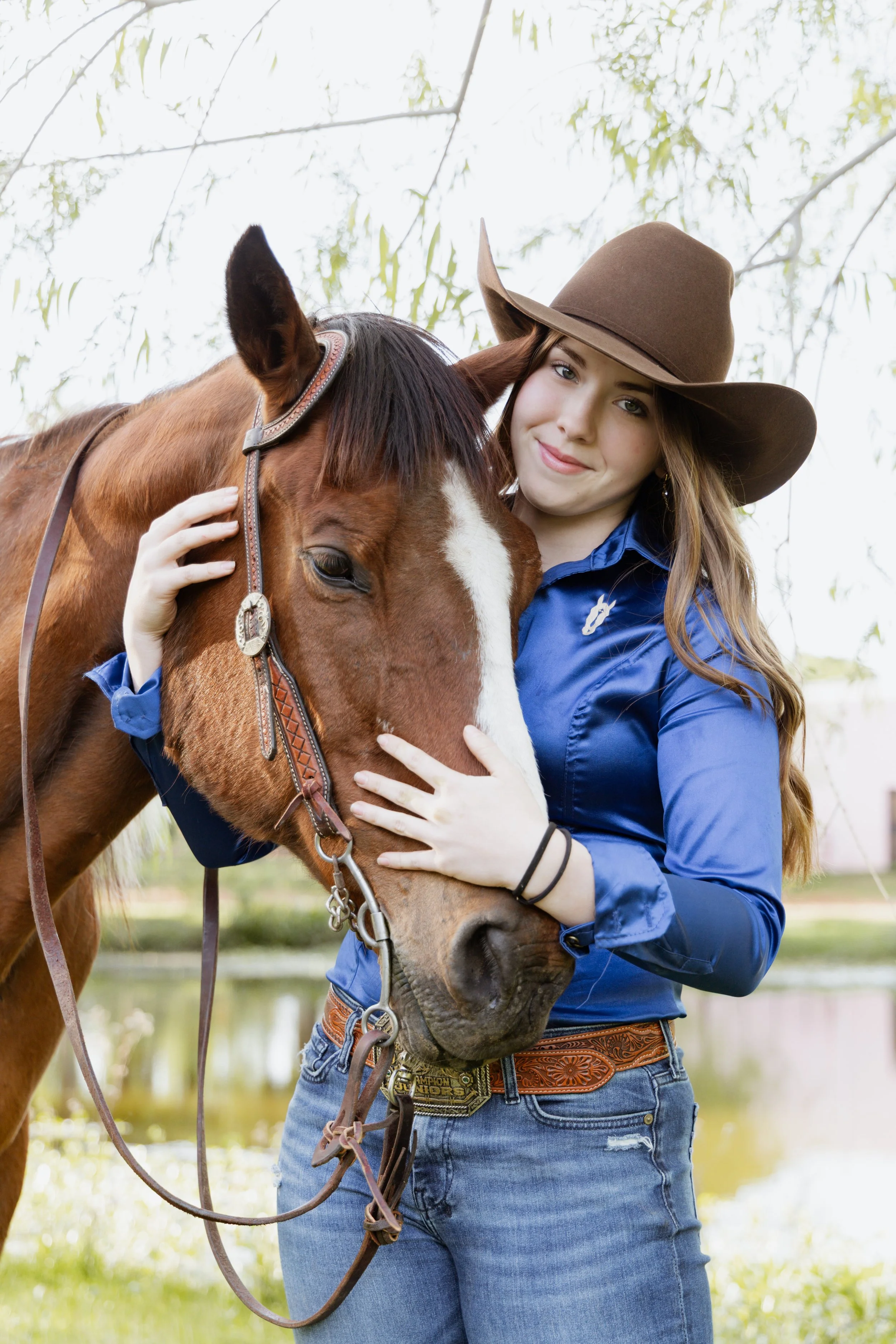 A young woman with long brown hair, wearing a brown wide-brimmed hat and a blue shirt, is gently holding a brown and white horse's face, standing outdoors with trees and a pond in the background.