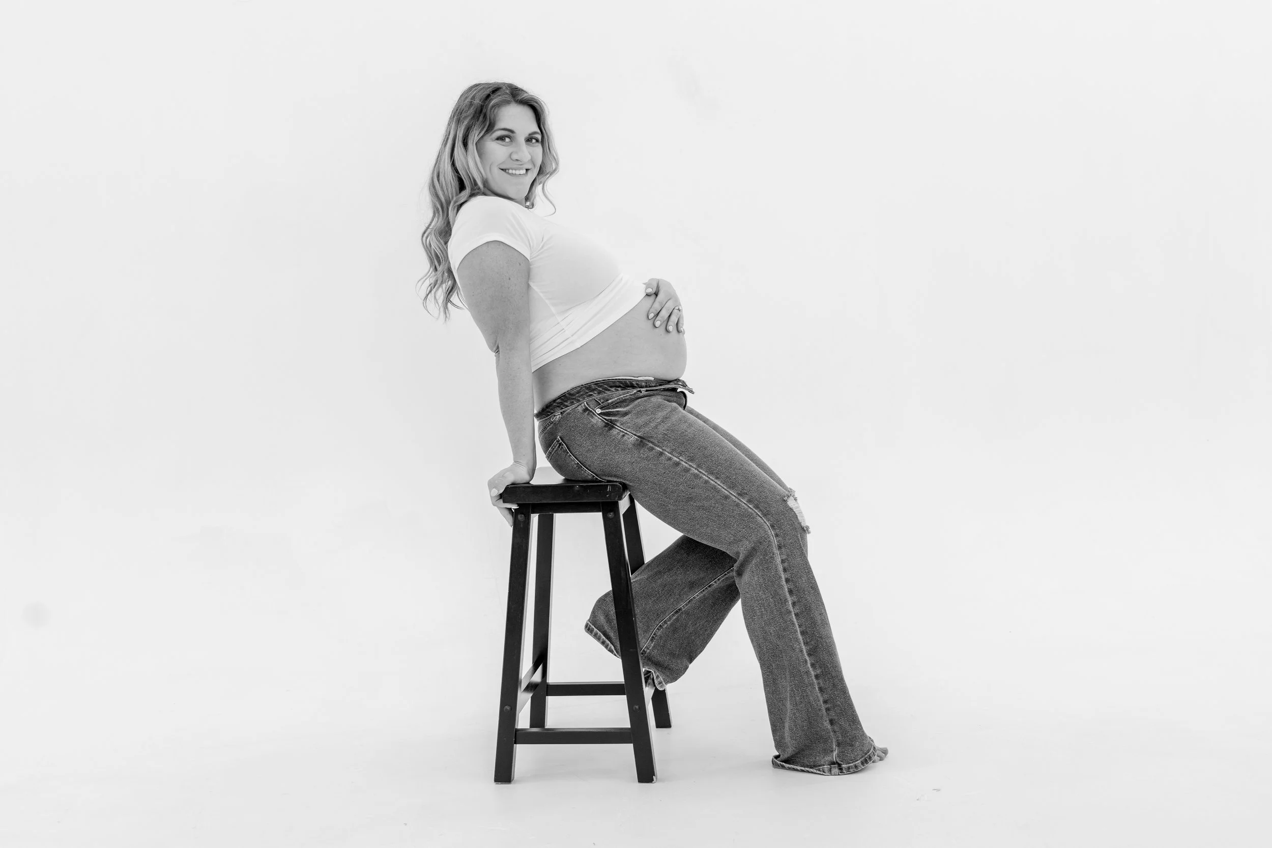 A smiling woman with wavy hair sitting sideways on a stool, wearing a white t-shirt and jeans, with her belly exposed, against a plain white background.