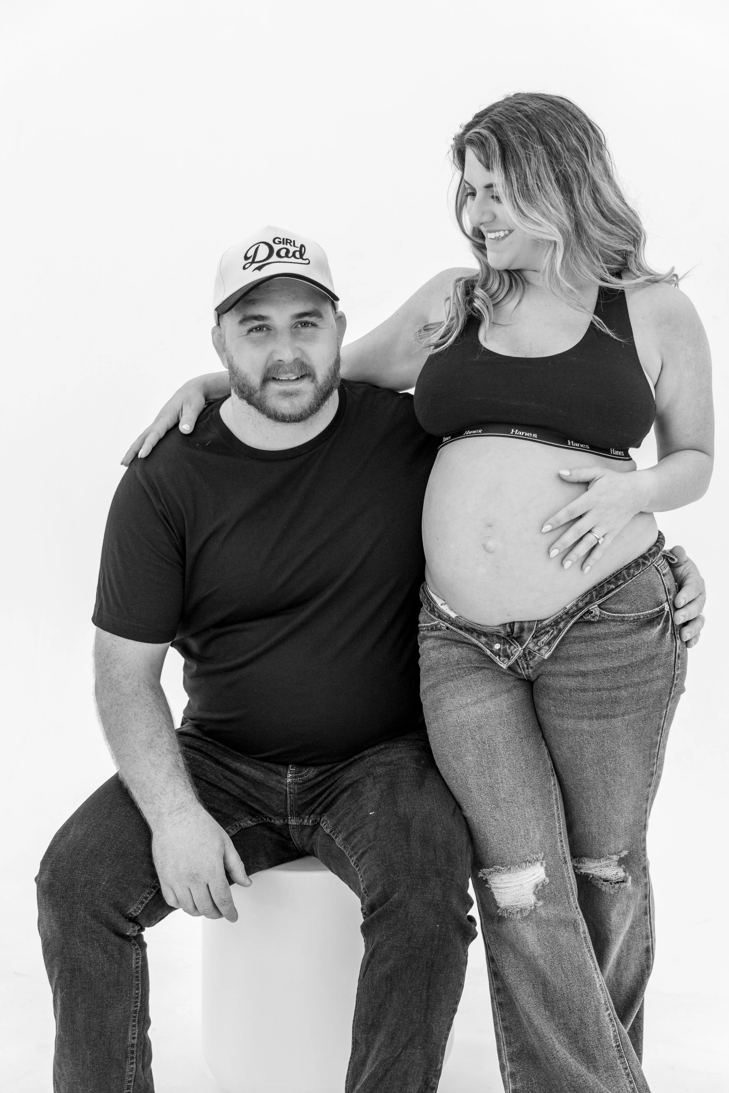 A woman with long, wavy hair and a pregnant belly wearing a black sports bra and ripped jeans. She is smiling and looking at a man with a beard, wearing a cap that says 'Girl Dad' and a black T-shirt. The man is seated on a white stool, and the woman